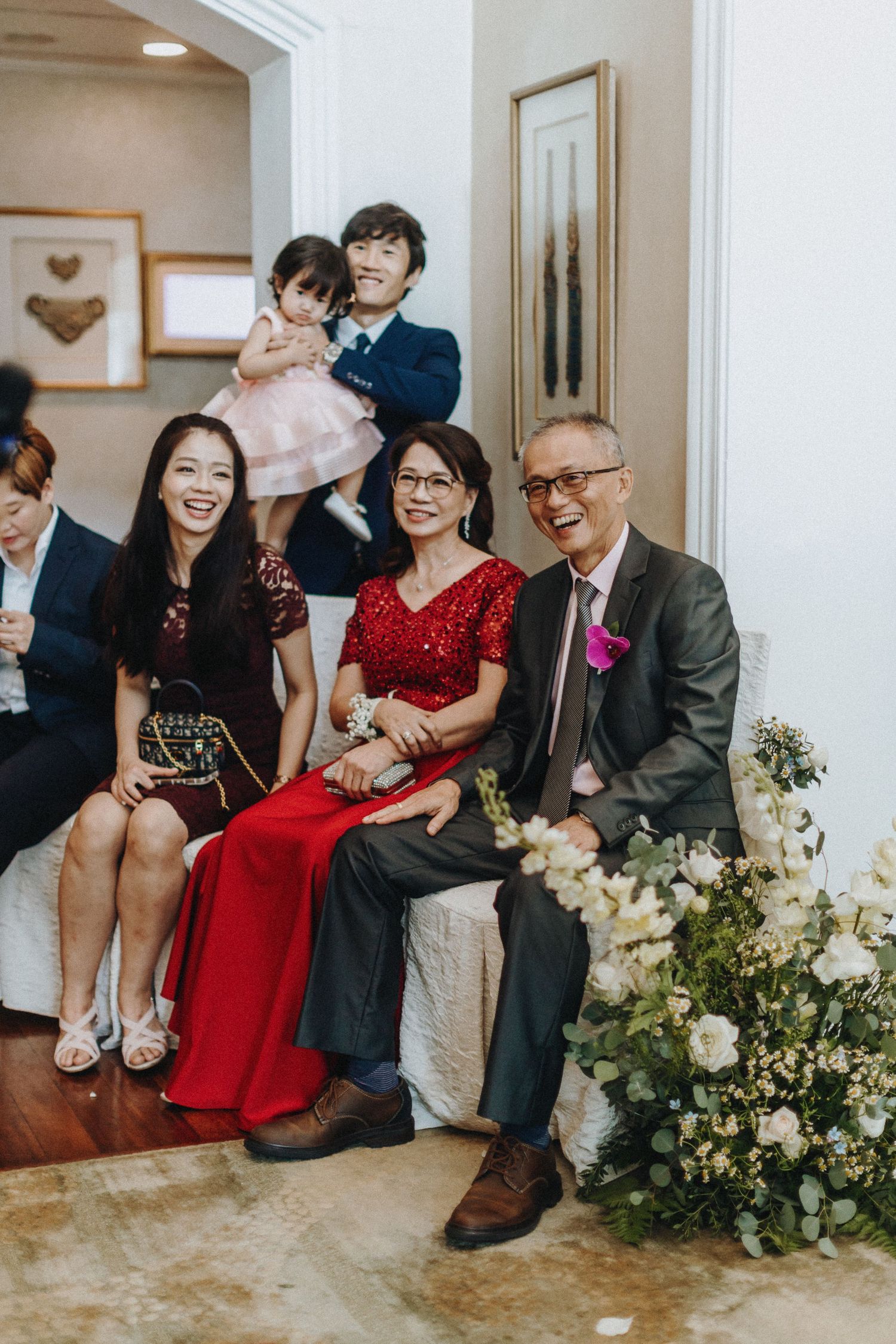 A family gathering with people sitting on a couch near floral arrangements at a special occasion.