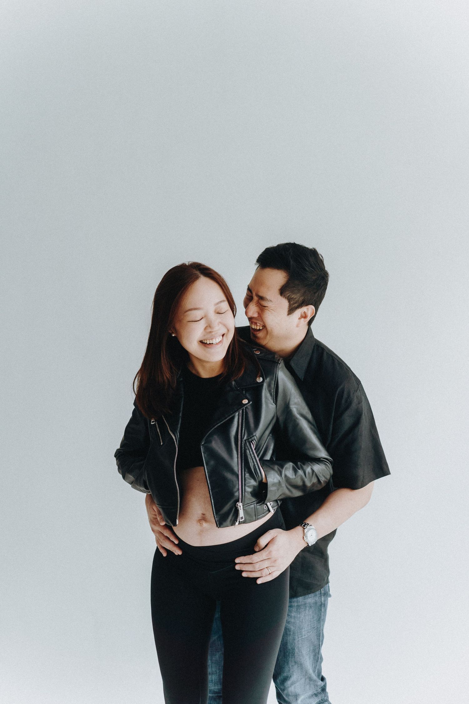 A candid moment between a couple wearing black outfits against a light grey background.