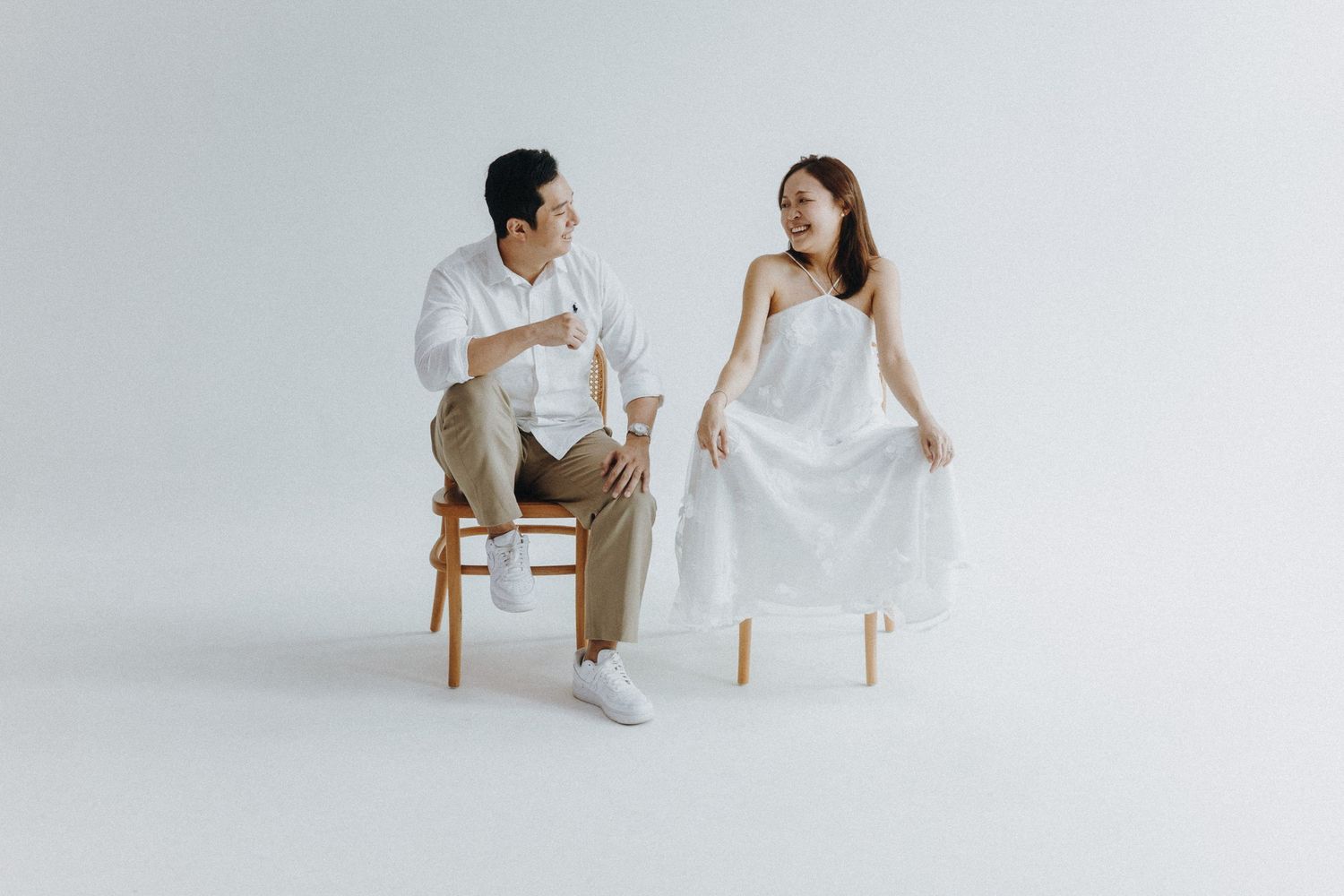 Couple in white attire seated on wooden chairs having an intimate conversation against a plain backdrop.