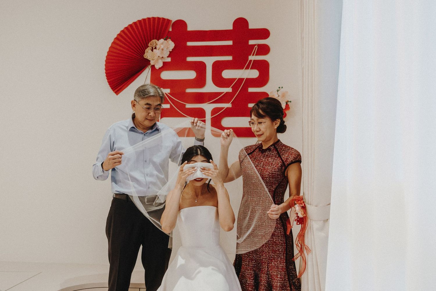 A joyful wedding celebration scene against a white wall with red Chinese double happiness symbol and paper fan decoration.