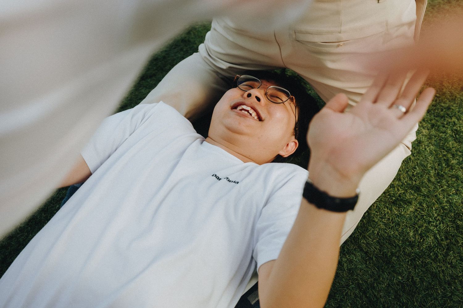 Person lying on grass wearing a white t-shirt and smartwatch while laughing.