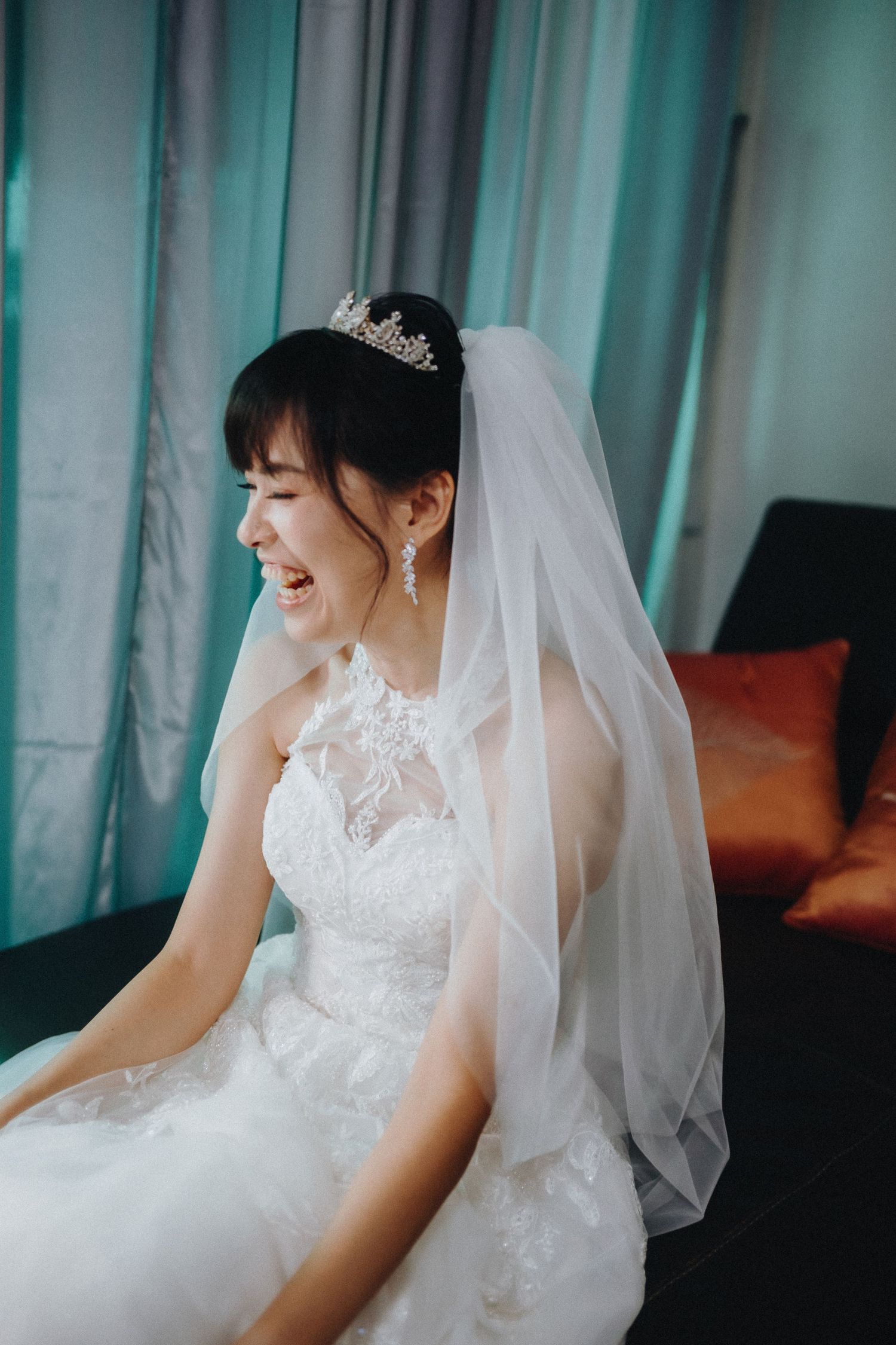 A bride in a white lace wedding gown and veil smiles while sitting indoors.