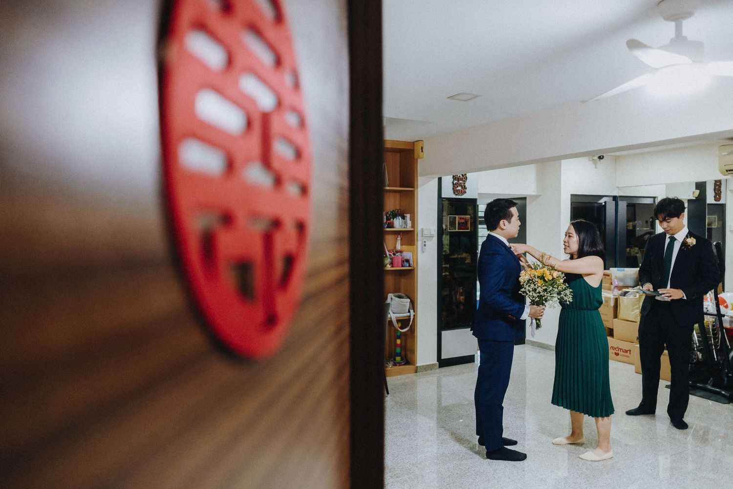 Three people stand in conversation in a bright interior room with a red decorative wall piece visible in the foreground.