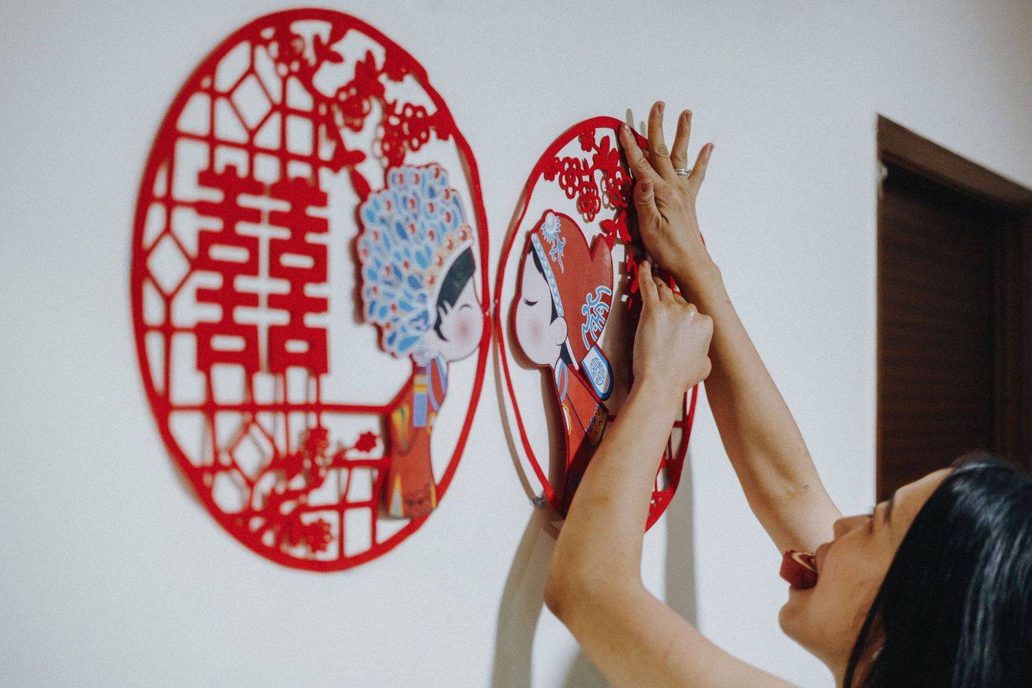 Someone hanging decorative red double happiness paper cutouts on a white wall for Chinese wedding celebration traditions.