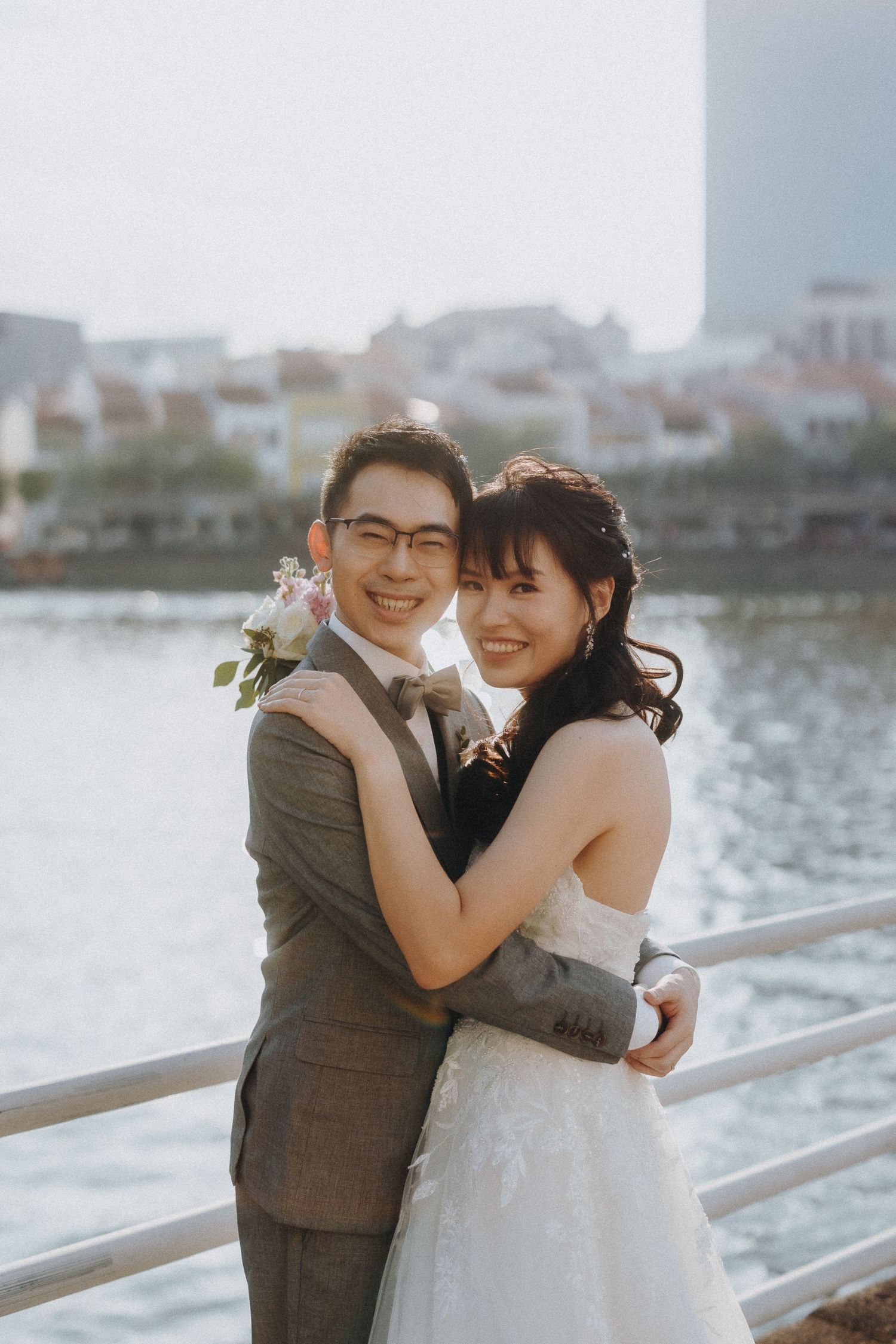 A newlywed couple embraces by a waterfront railing during their wedding photoshoot.