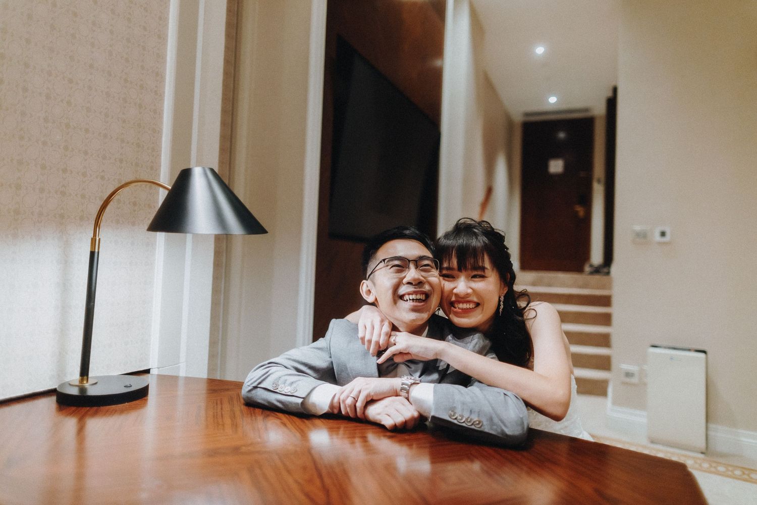 Two people share a joyful moment while leaning on a wooden desk with a modern lamp.