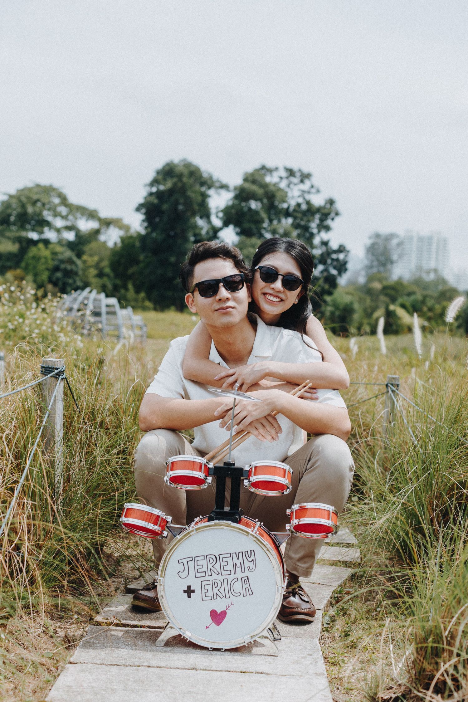 Couple wearing sunglasses posing with DIY drum set made from red and white containers in a field.