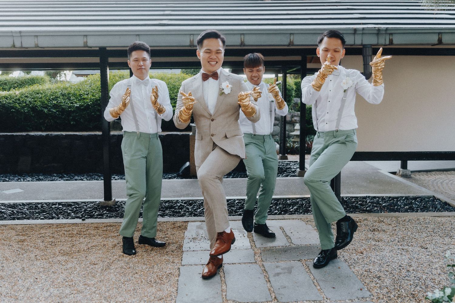 Four groomsmen in sage green pants and suspenders pose playfully outside a building.