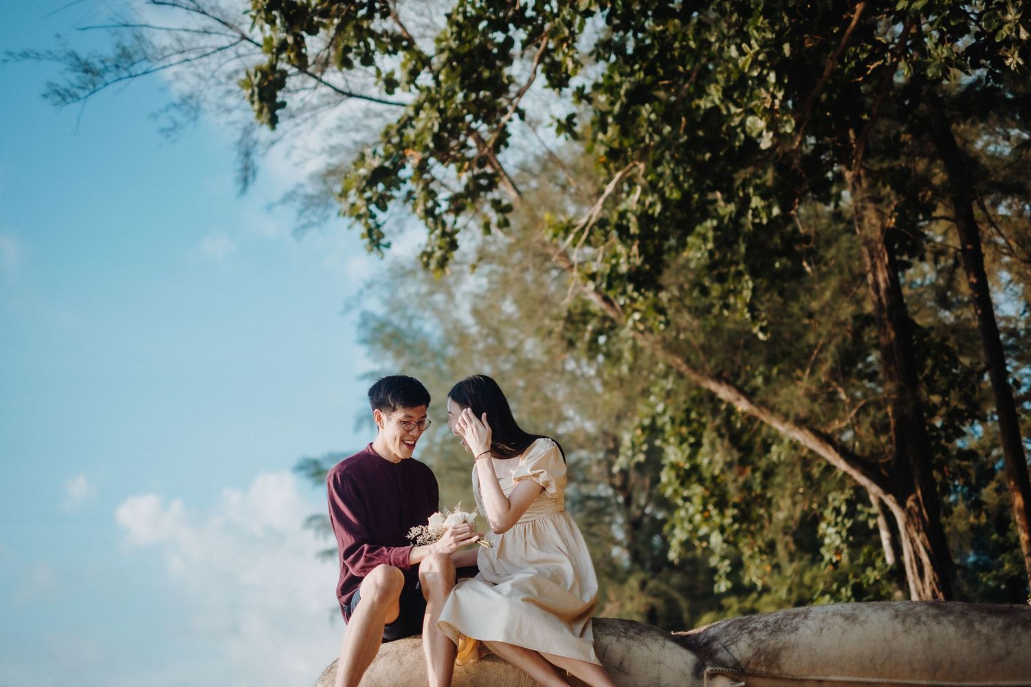 A romantic couple sits together on rocks against a bright blue sky with leafy trees framing the scene.
