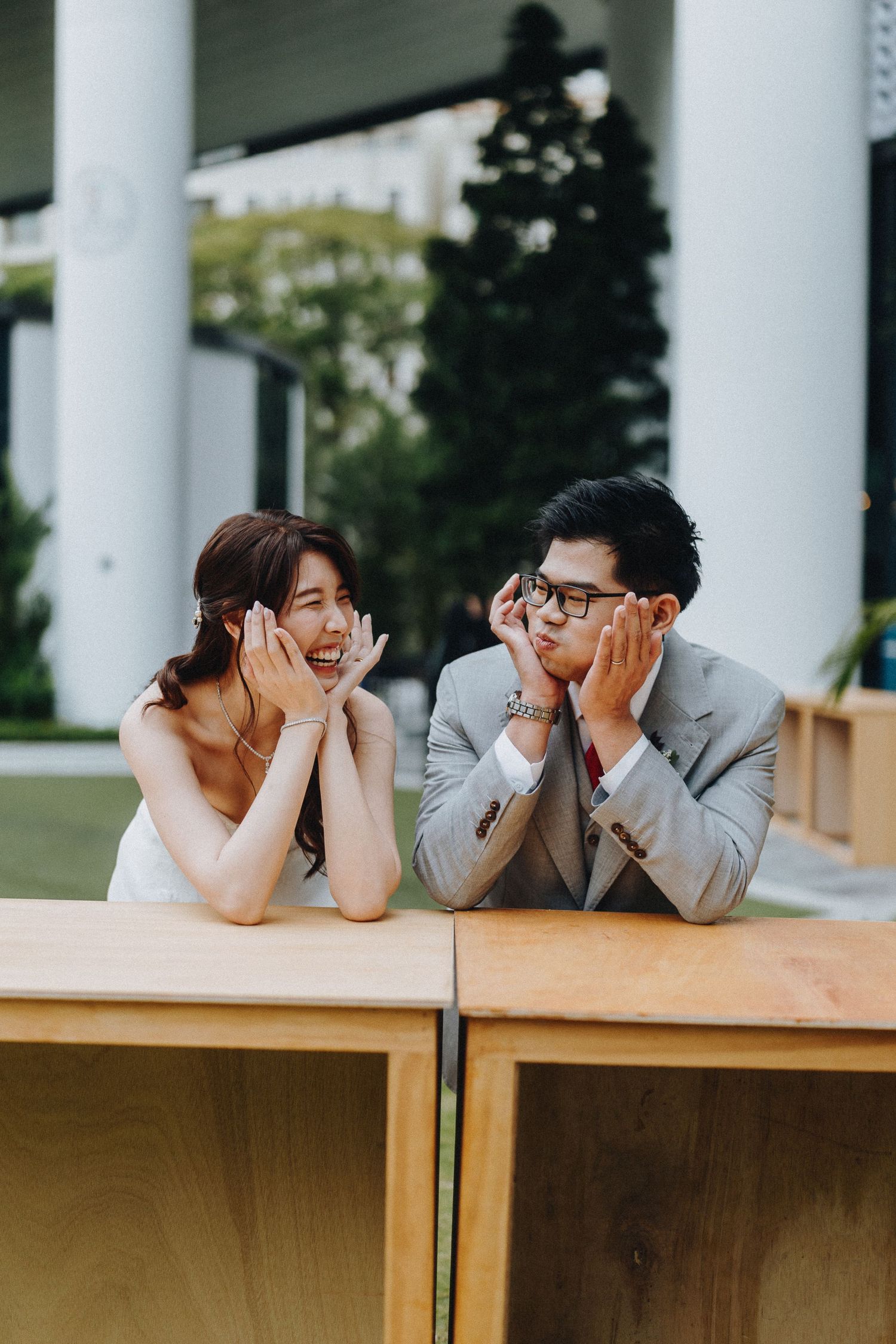 A couple leans on wooden tables outdoors while playfully resting their heads in their hands with joyful expressions.