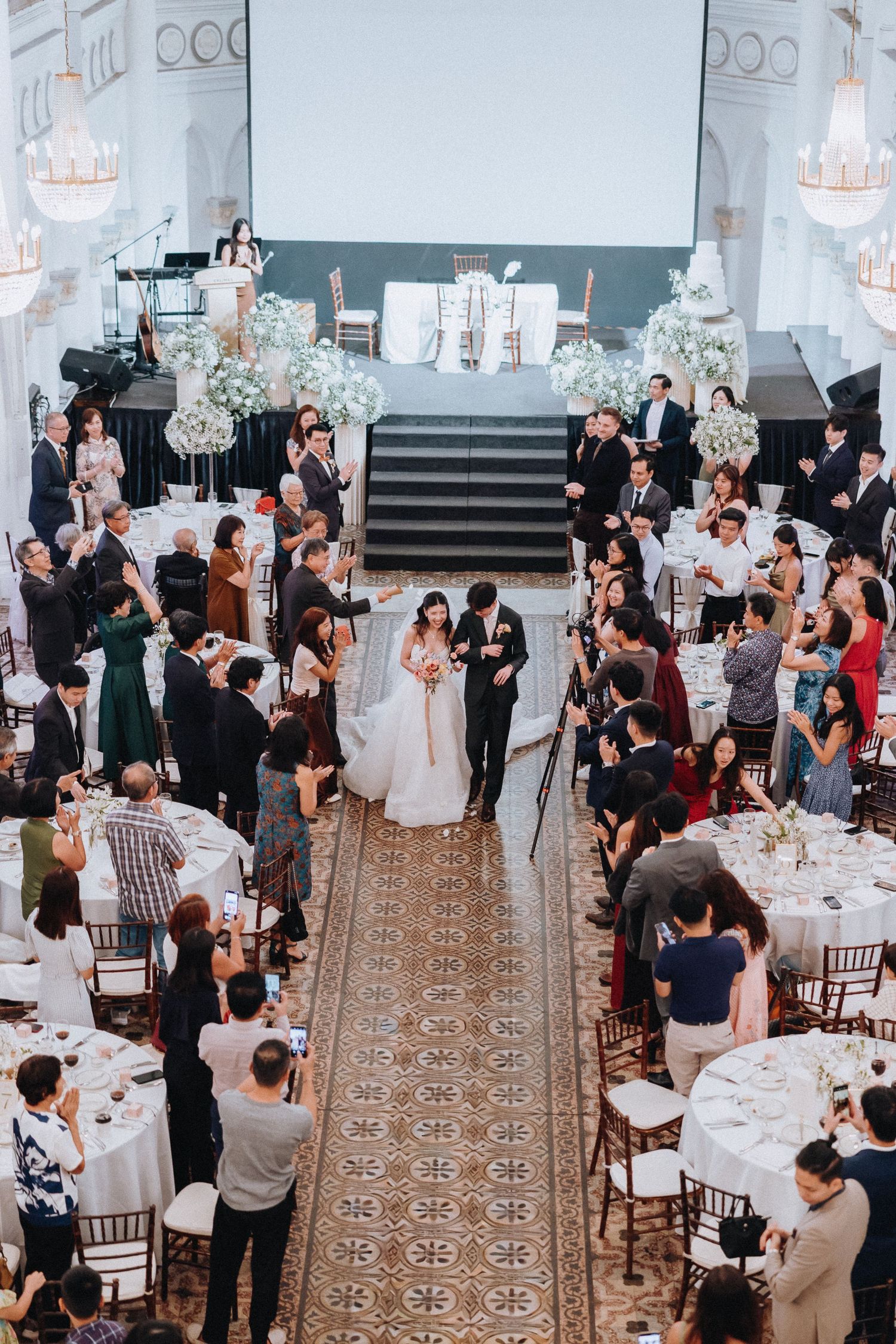 Aerial view of a wedding reception with bride and groom walking the aisle surrounded by guests in an elegant ballroom.