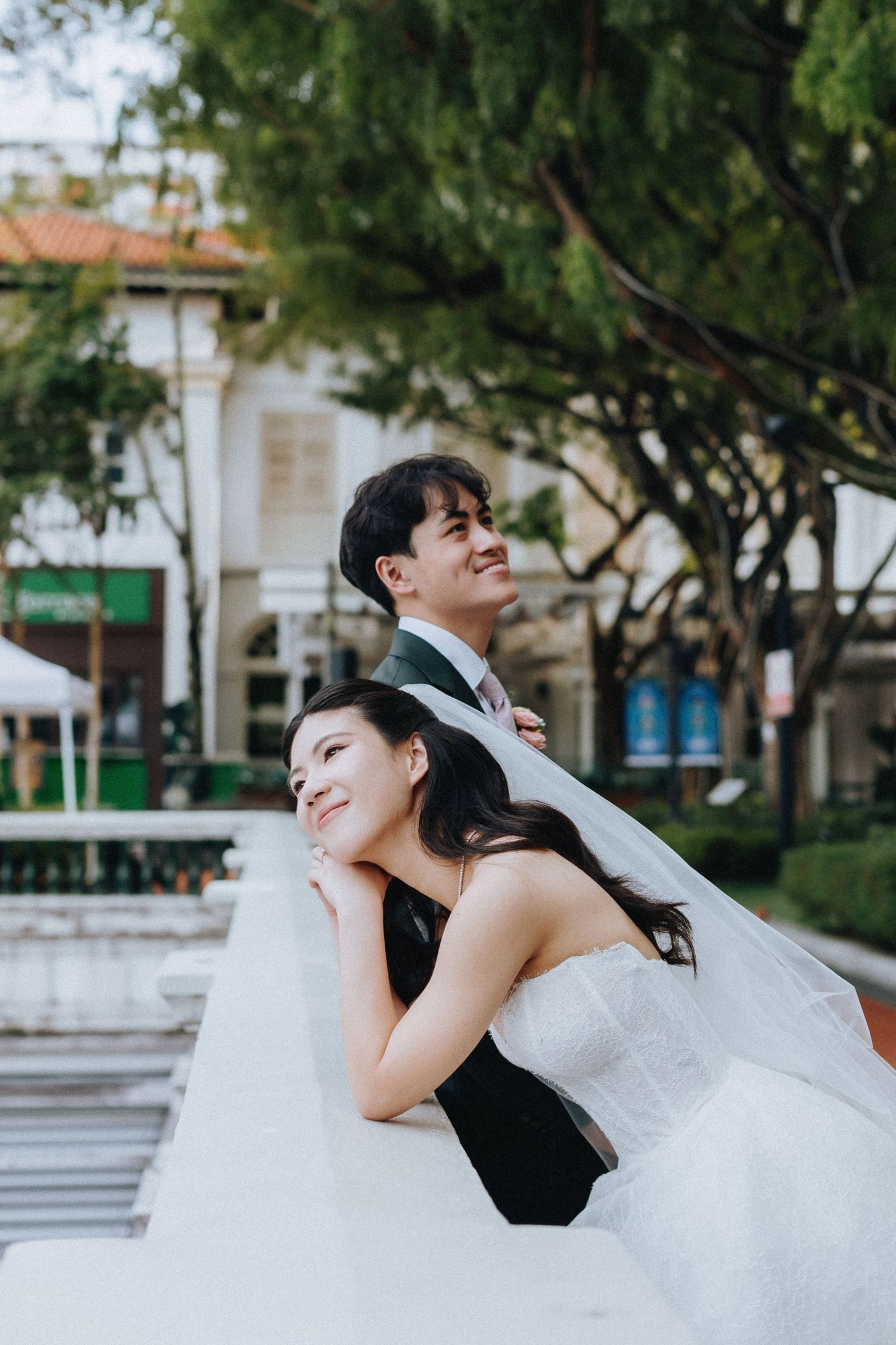 Bride and groom leaning on white ledge outdoors beneath green trees in a relaxed candid pose.