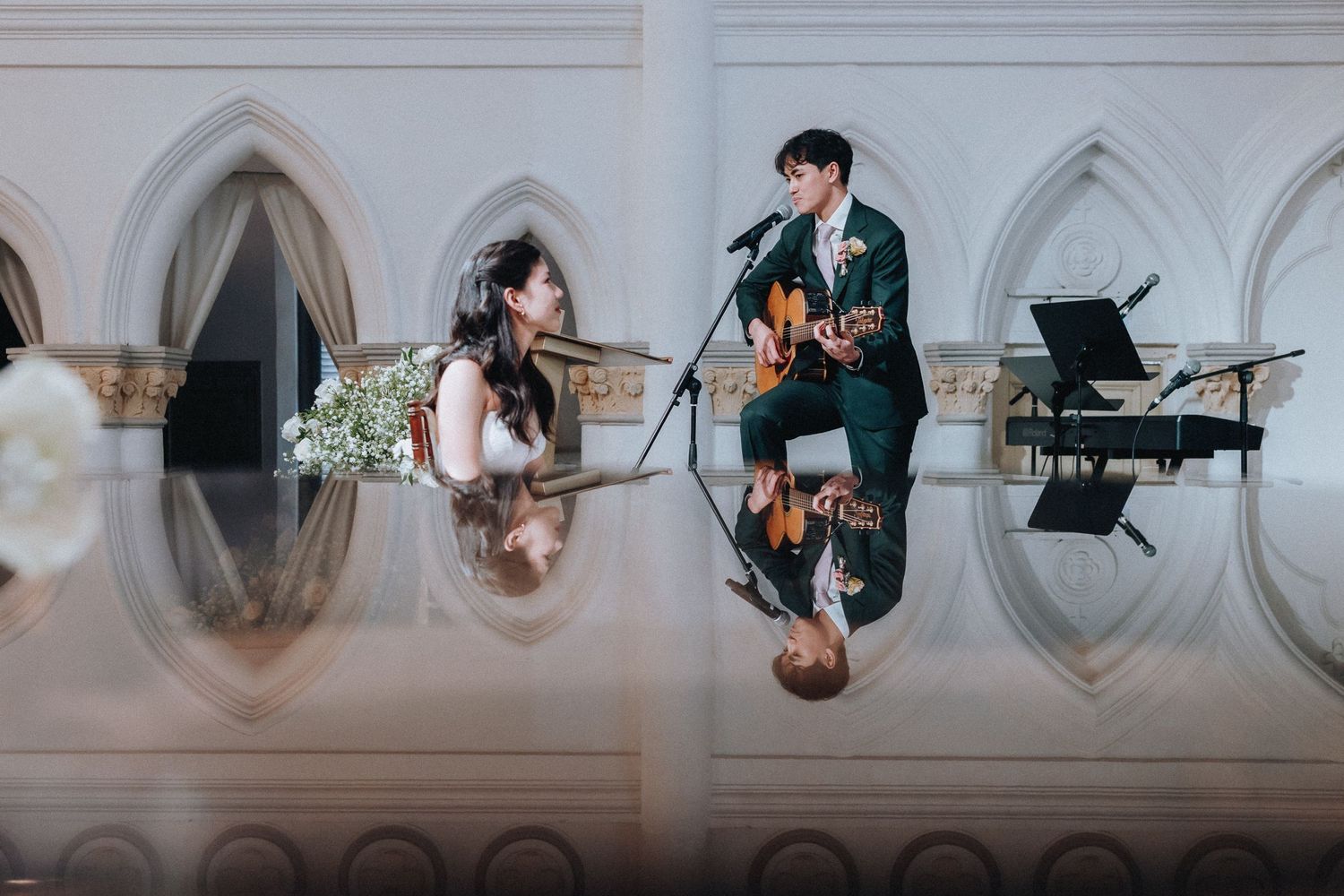 A couple performs music together at an elegant venue, man playing guitar at microphone while woman sits nearby with flowers.