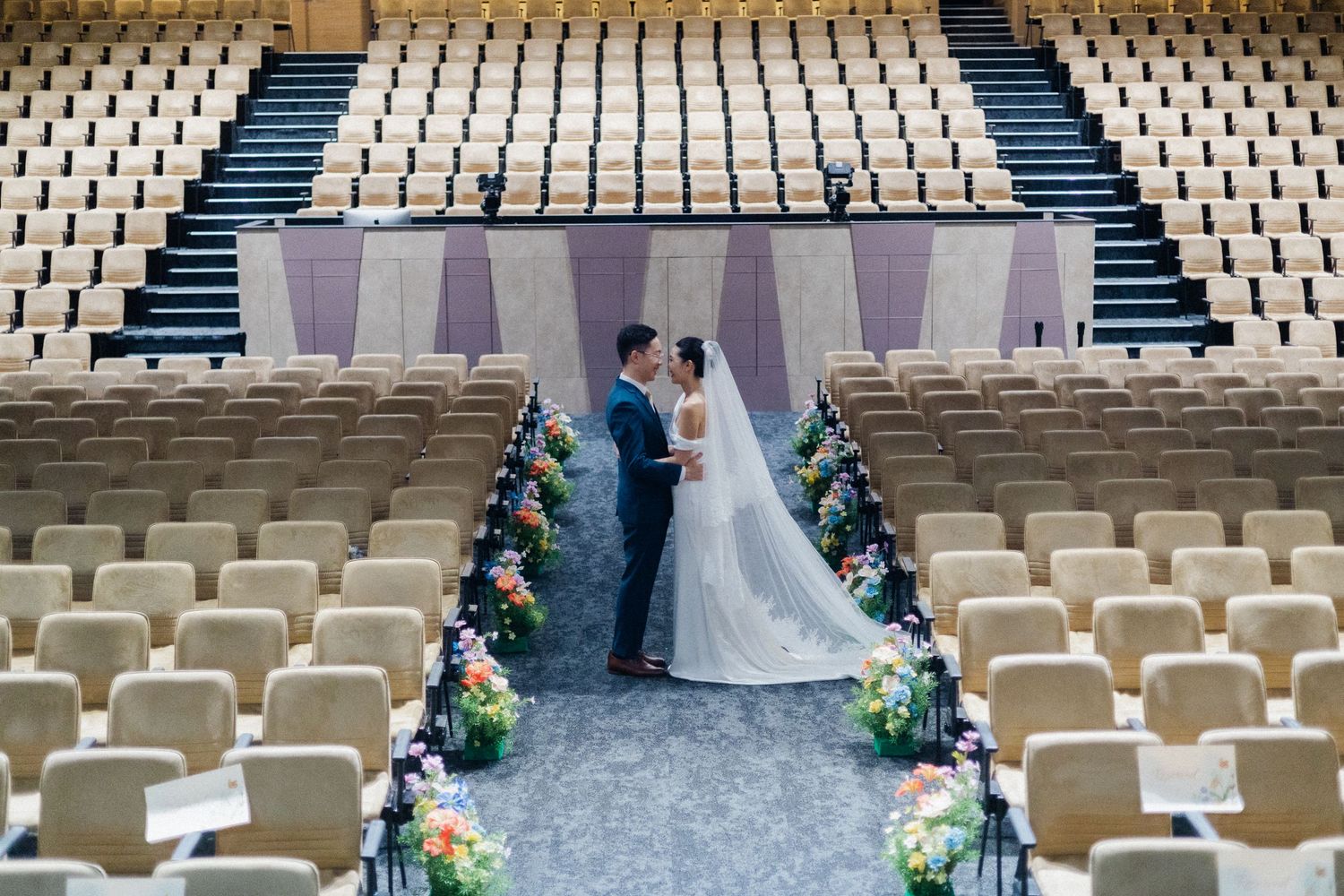 Wedding couple stands at the altar of an empty auditorium lined with colorful floral arrangements.