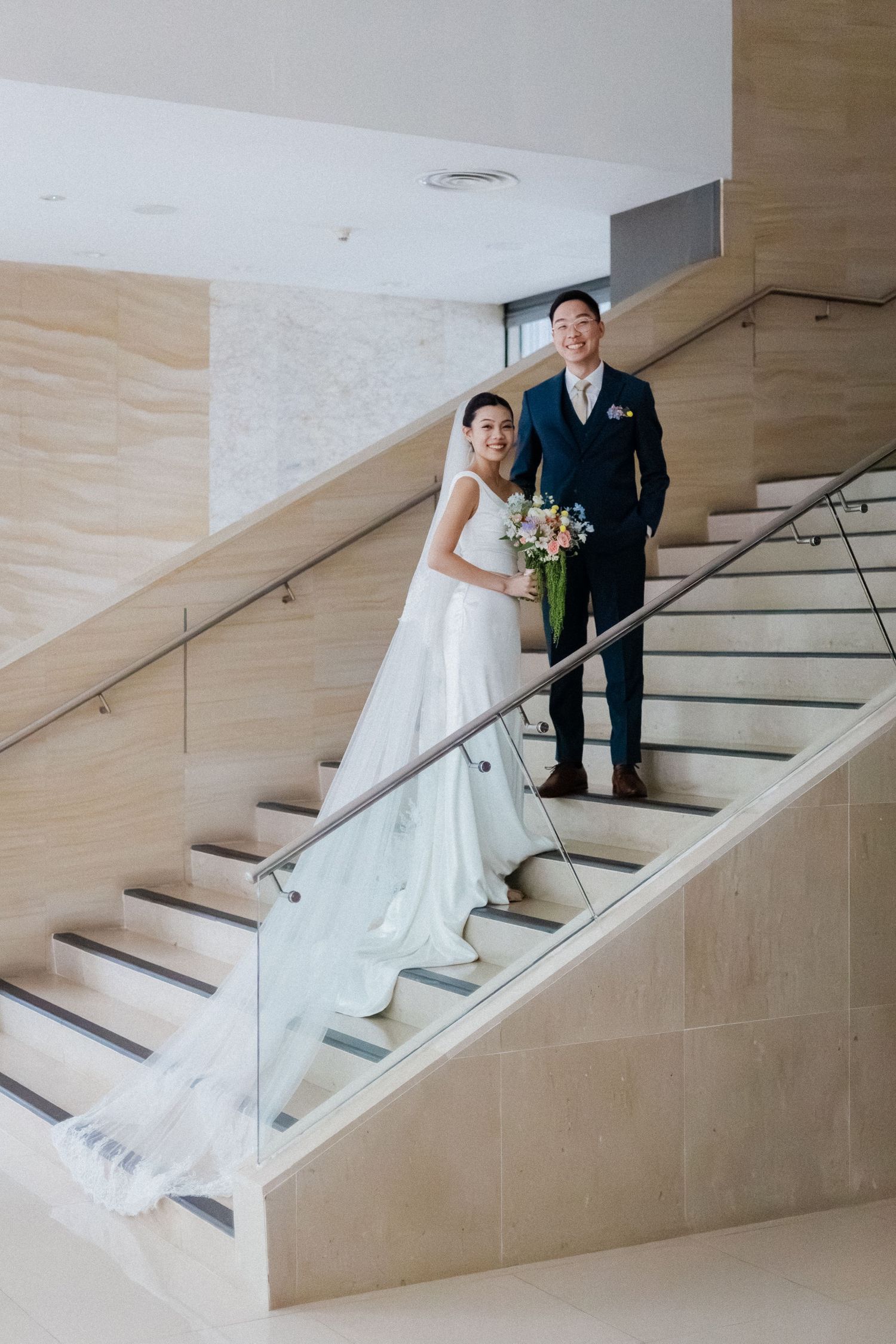 Asian bride and groom posing elegantly on modern staircase, bride in white gown with long veil, groom in navy suit holding bouquet.