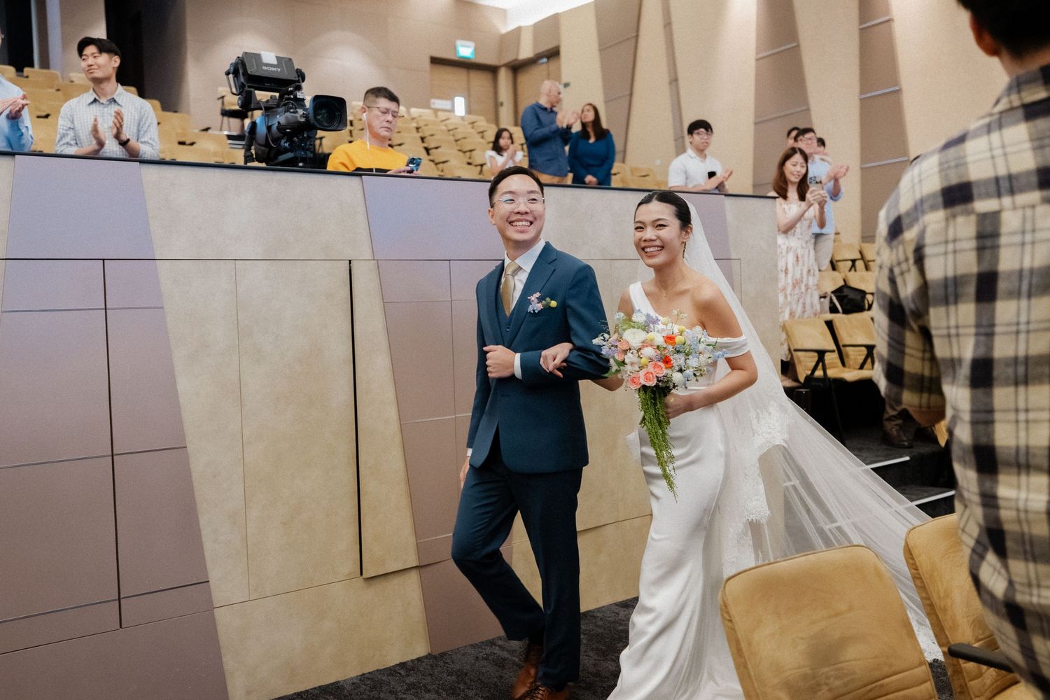 Joyful bride and groom walking down the aisle together in a modern auditorium ceremony venue.