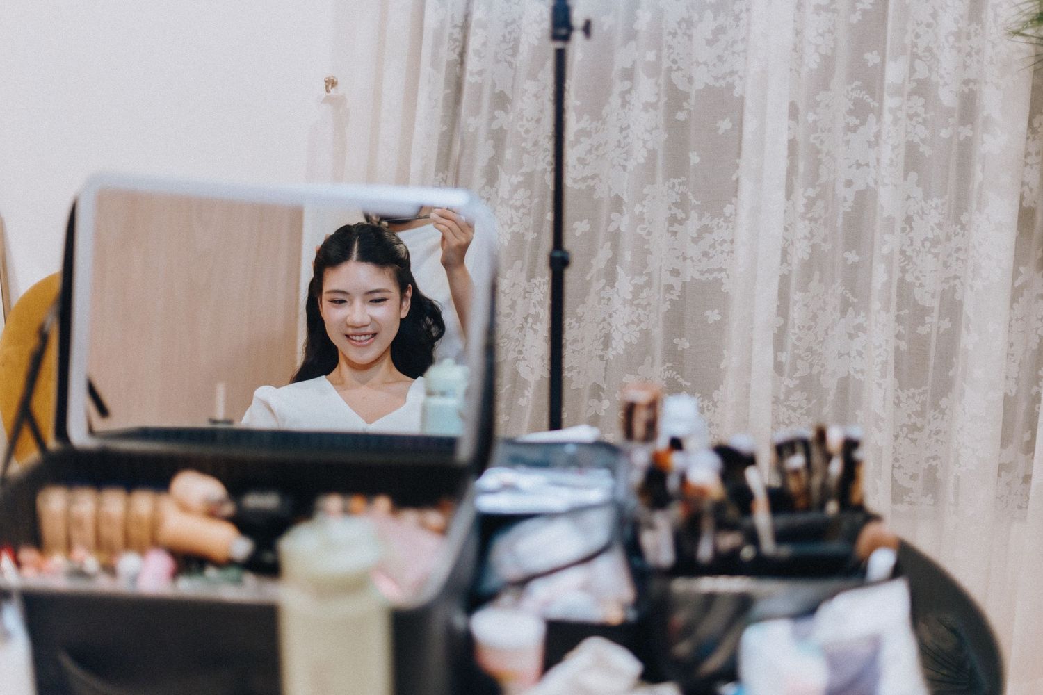 Young woman smiling while styling her hair in a mirror surrounded by makeup brushes and beauty products on a vanity.
