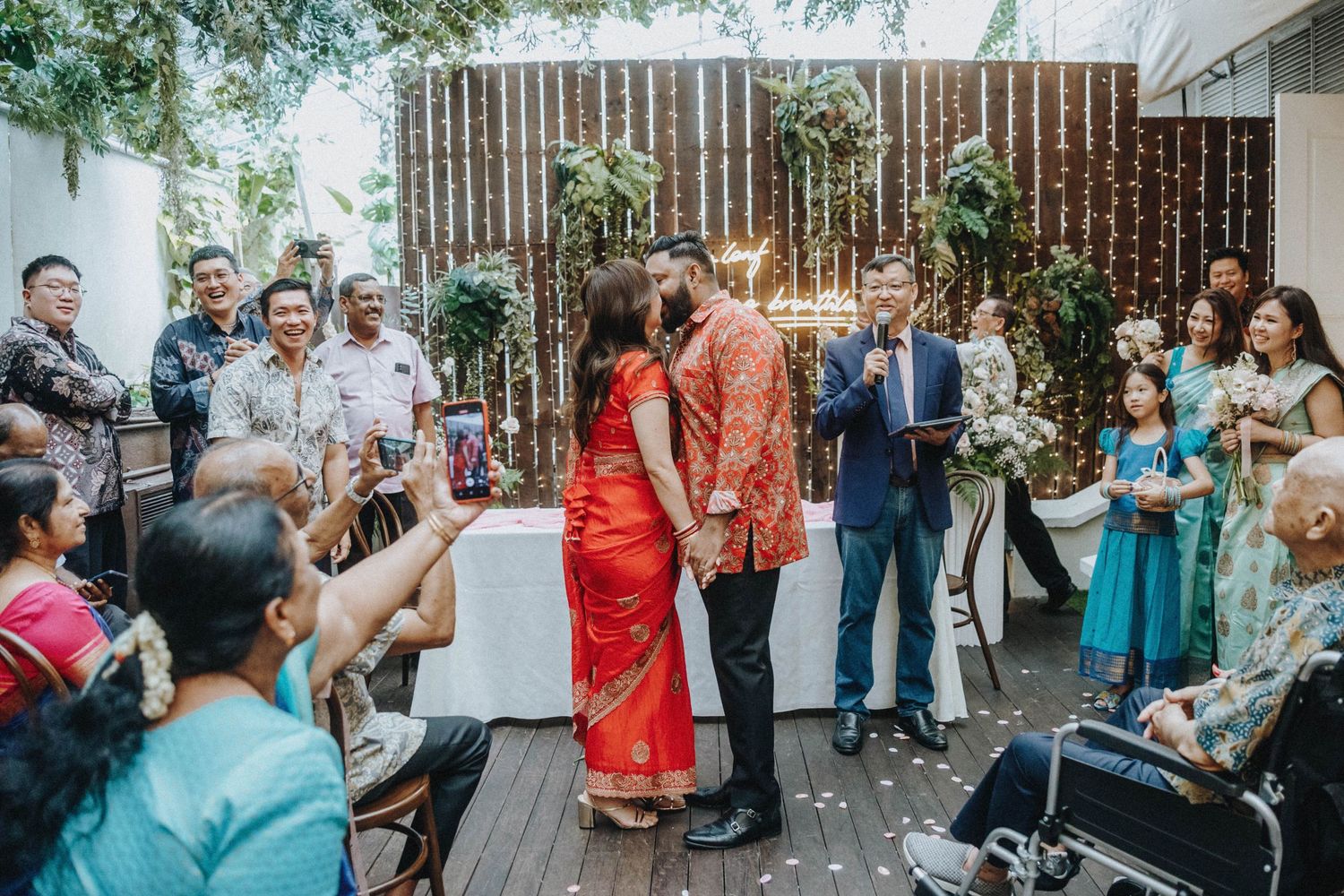 A couple shares a kiss surrounded by guests at an outdoor multicultural wedding reception.