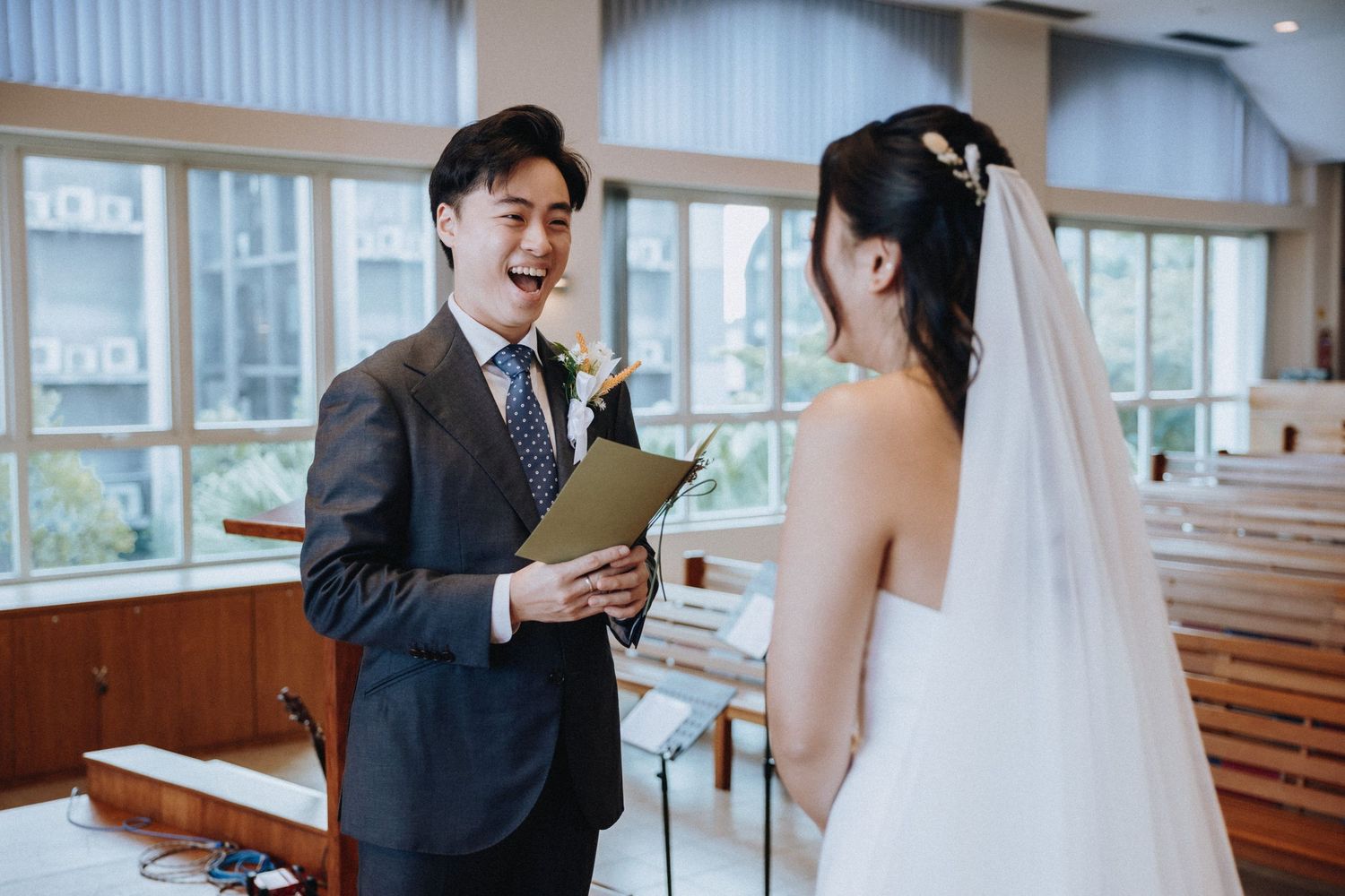Groom laughing joyfully while reading wedding vows to bride in white dress and veil during indoor ceremony.