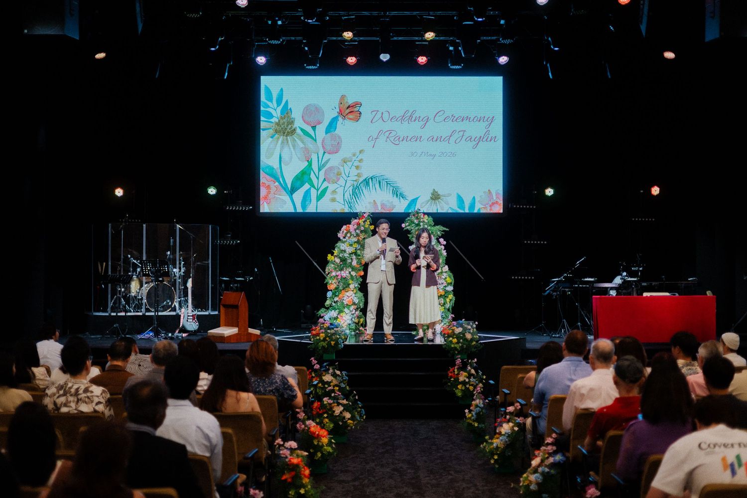 A wedding ceremony on stage with floral arch, couple and officiant, colorful tropical backdrop screen, and seated guests in attendance.