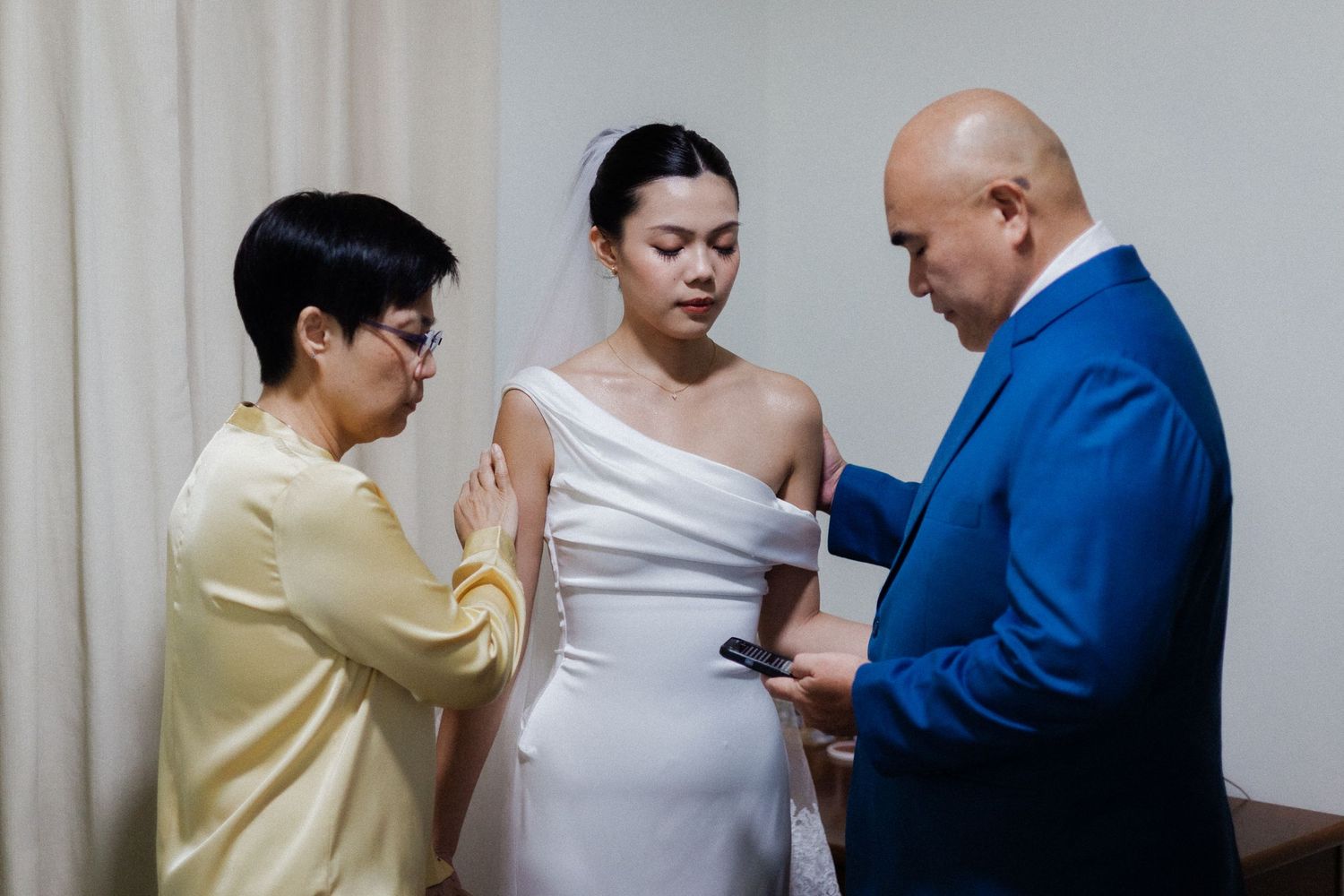 Bride in white one-shoulder gown stands with two guests in formal attire during a wedding preparation moment.
