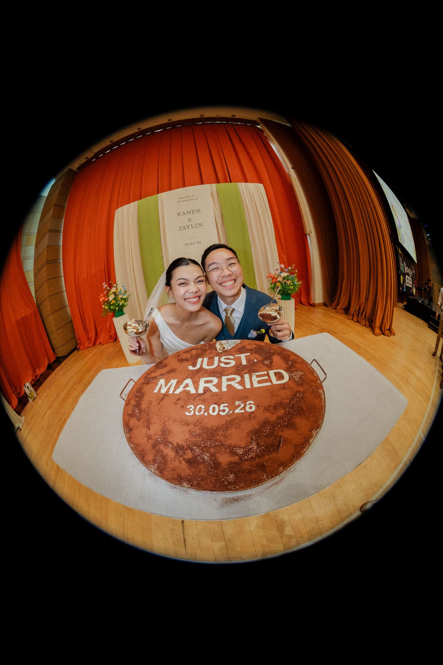 Fisheye lens photo of newlyweds posing behind Just Married cake on wooden floor with orange curtain backdrop.