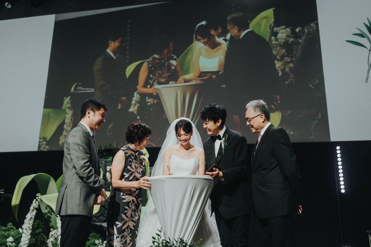 Group gathering around a table during wedding ceremony with projector screen in background.