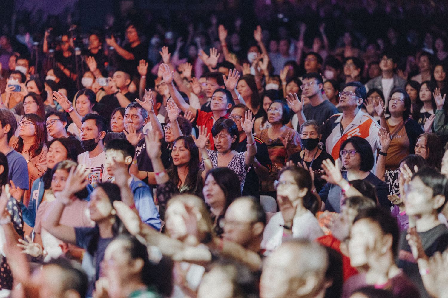 An energetic crowd of concert-goers raise their hands and phones during a live performance.