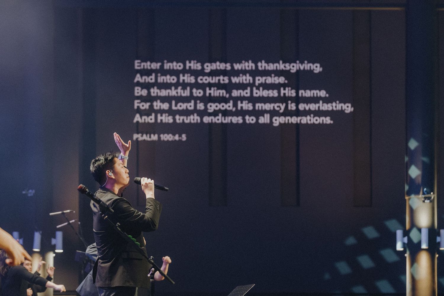 A worship stage setting with biblical text from Psalm 100:4 displayed on a screen above during a concert performance.