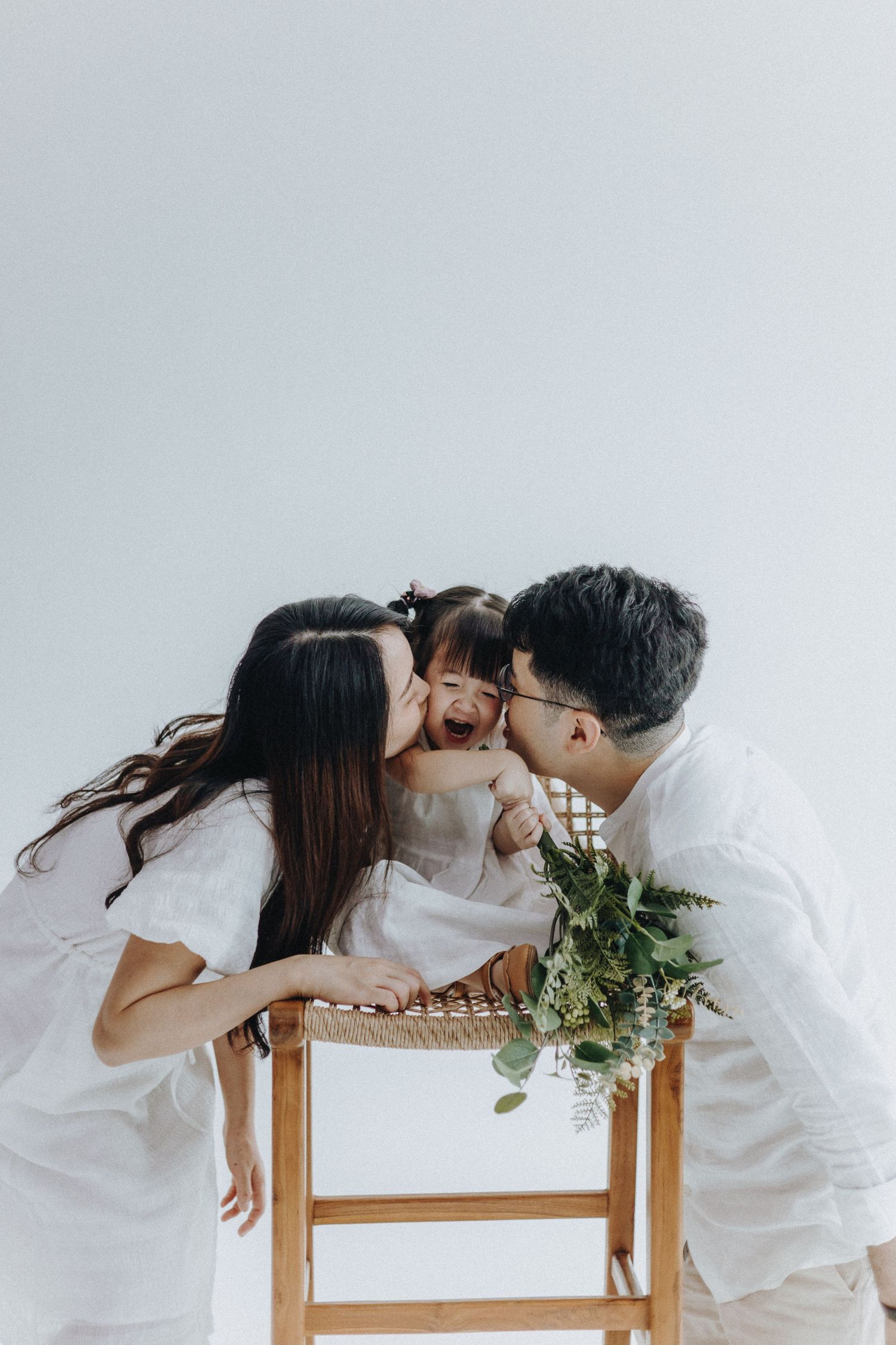 Couple in white clothing share intimate moment while resting on wooden chairs with greenery.