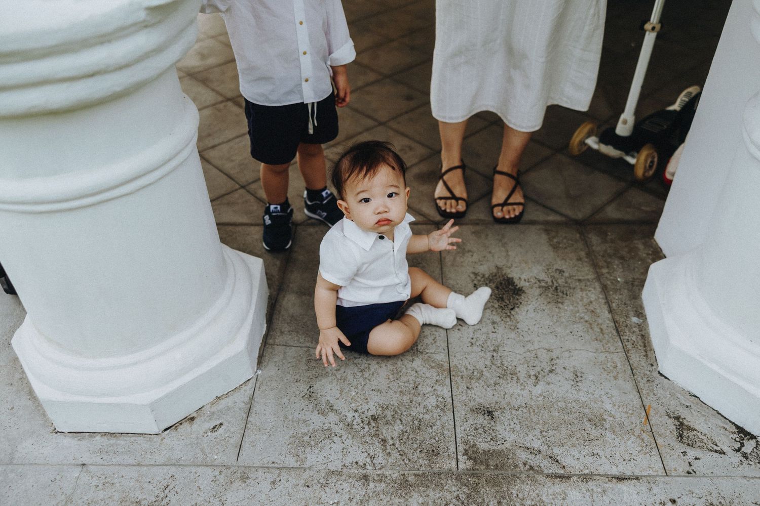 Small figure sits between white columns on tiled floor holding something white in their hands.
