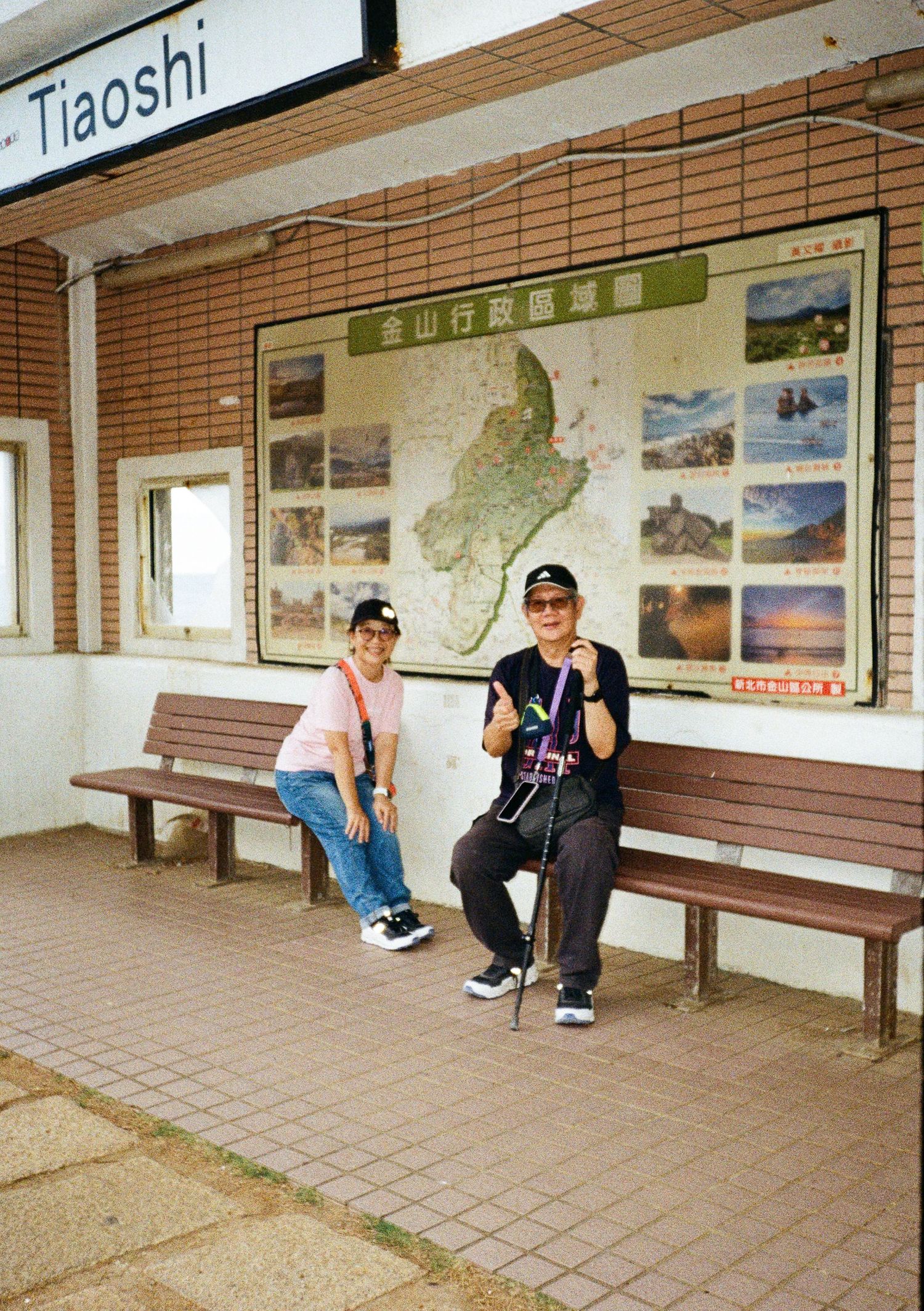 Train station waiting area with wooden benches and tourism photos displayed on tiled wall.