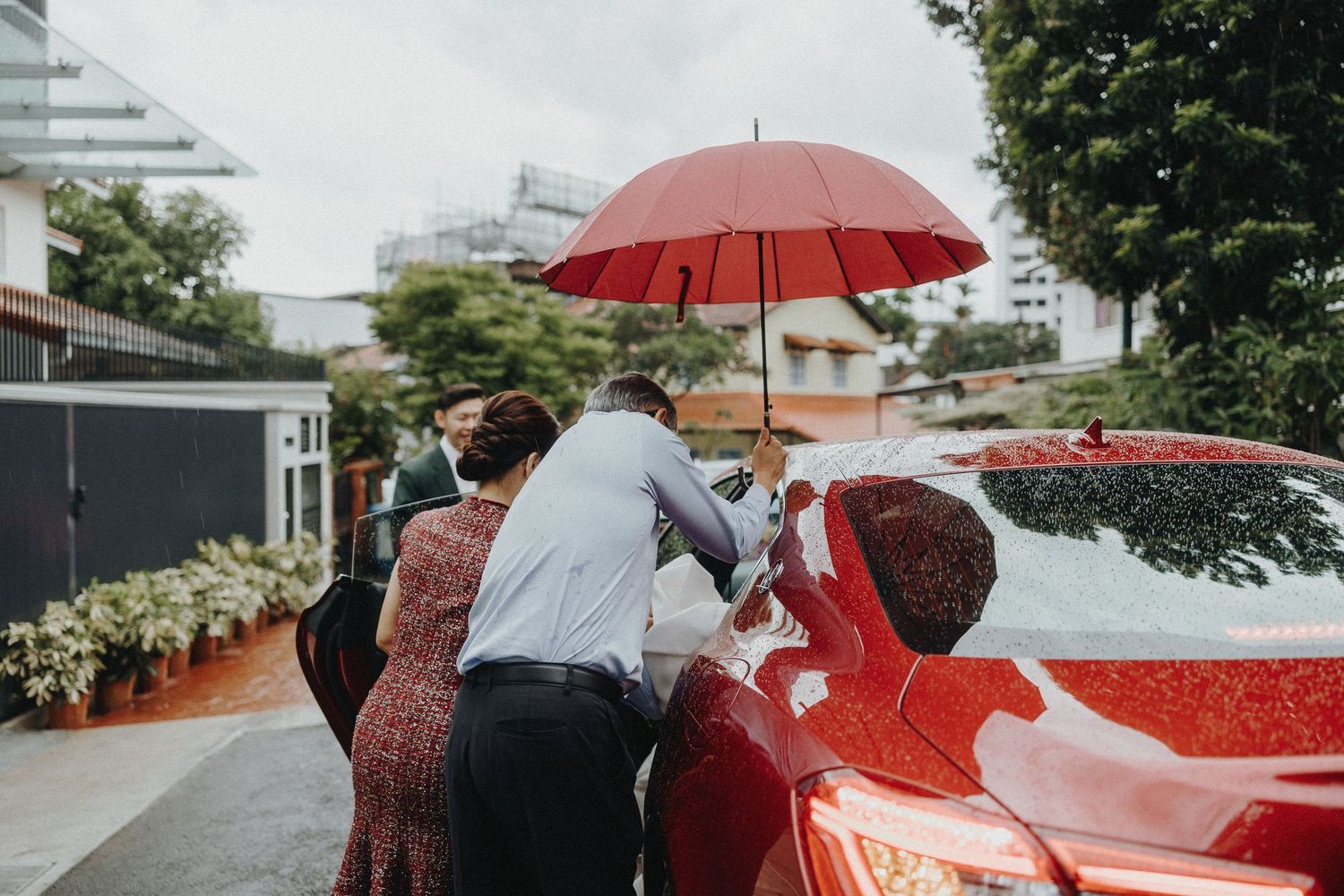 Wedding party walking outdoors holding red umbrellas under cloudy skies.