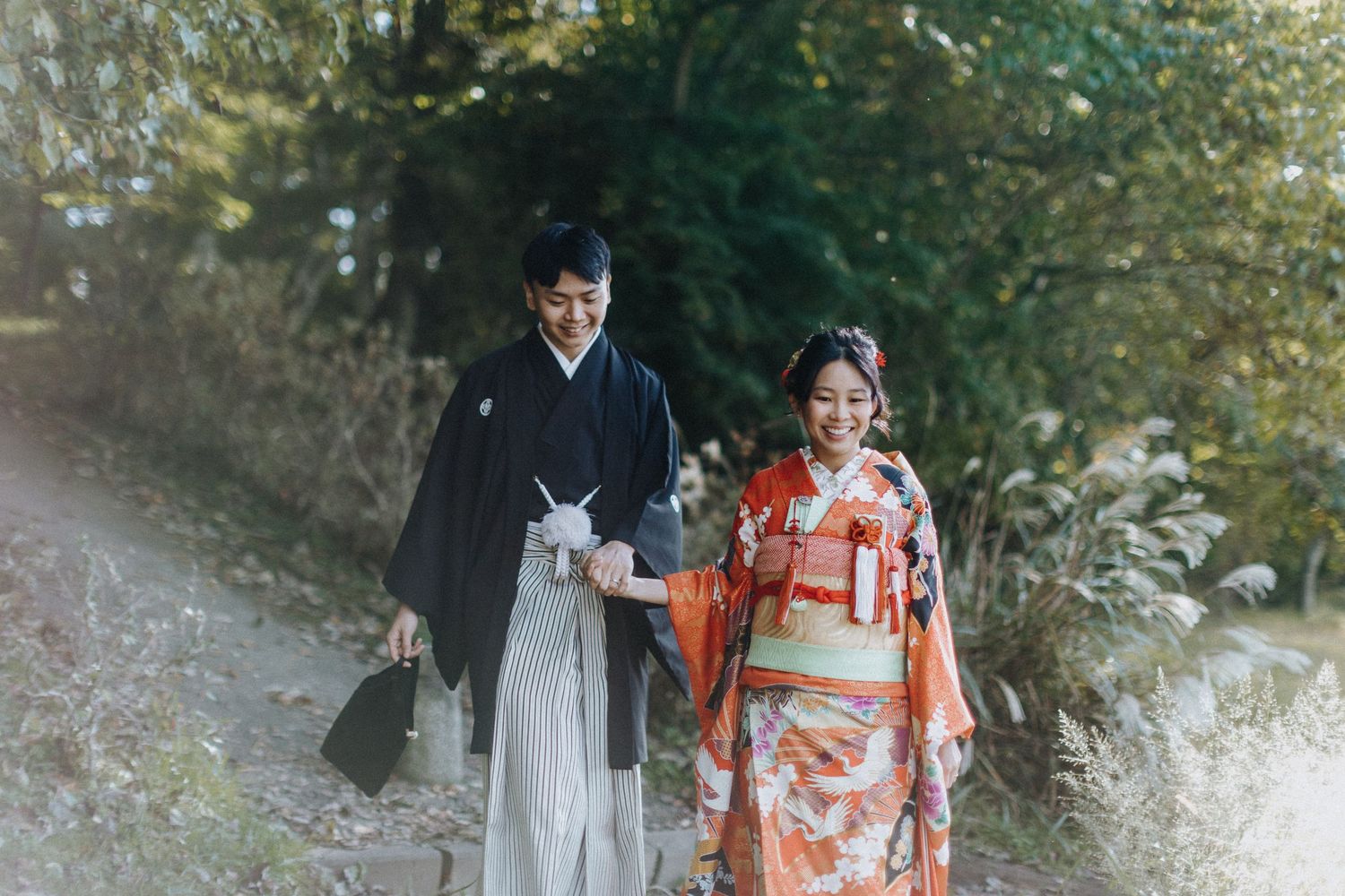 A couple in traditional wedding attire, with a kimono and white suit, walk together in a scenic outdoor setting.