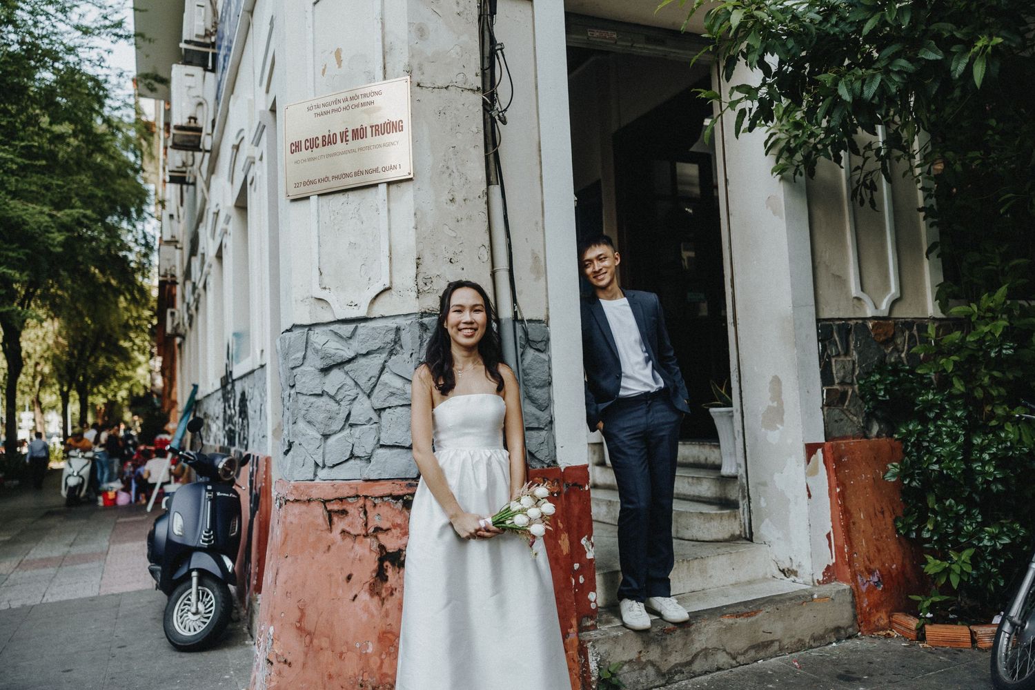 A person in a white wedding dress stands outside an urban building with rustic orange and gray walls and greenery.