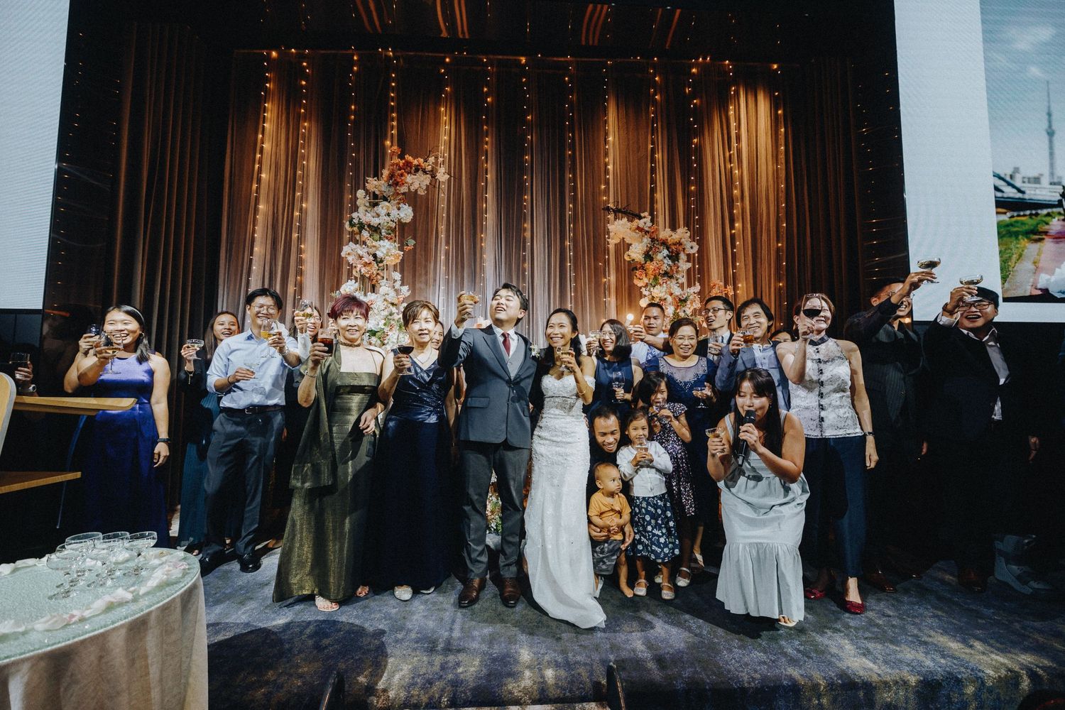 Large wedding party group photo with string lights and floral decorations in the background.