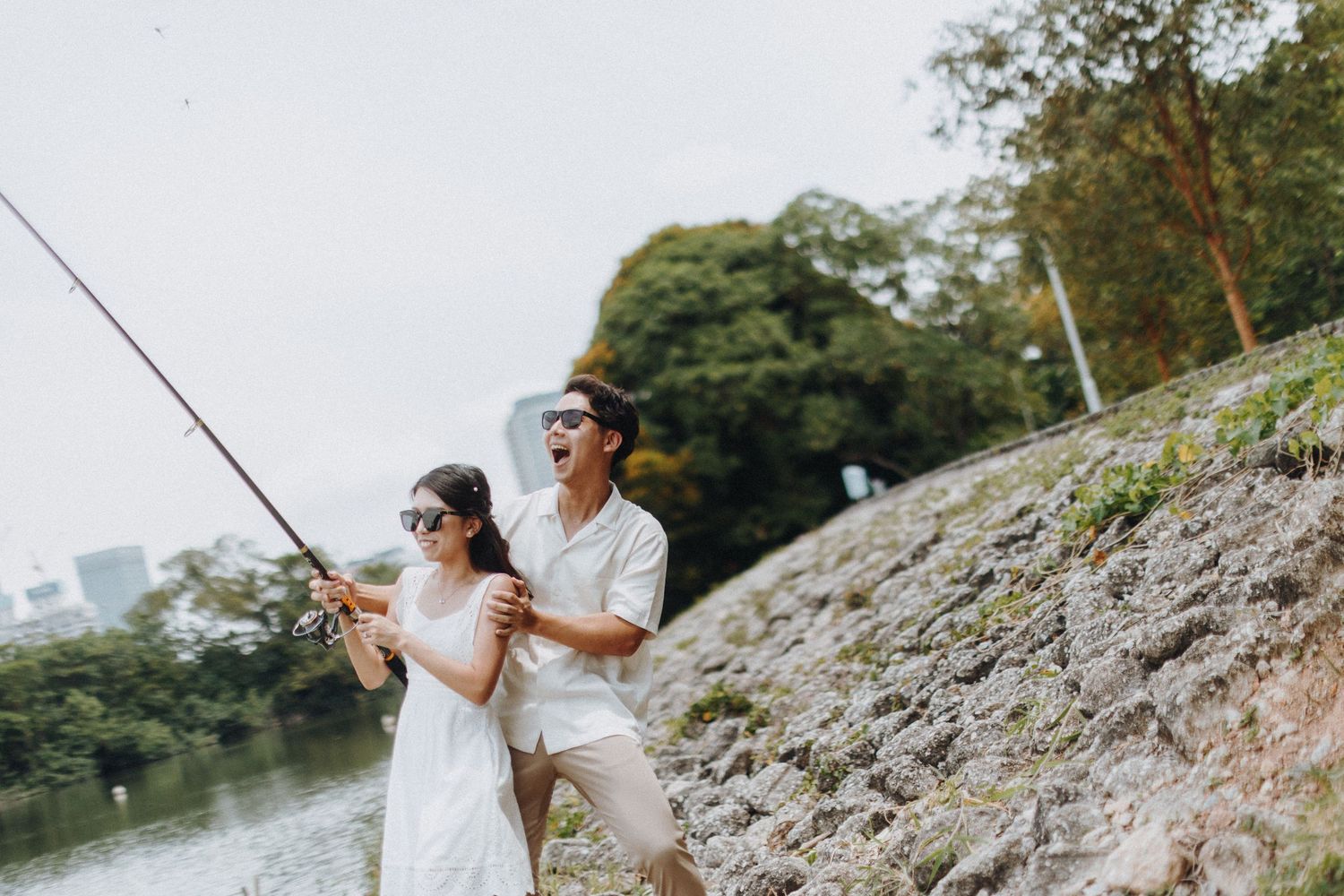 Two people enjoying fishing together on a rocky riverbank with trees in the background.