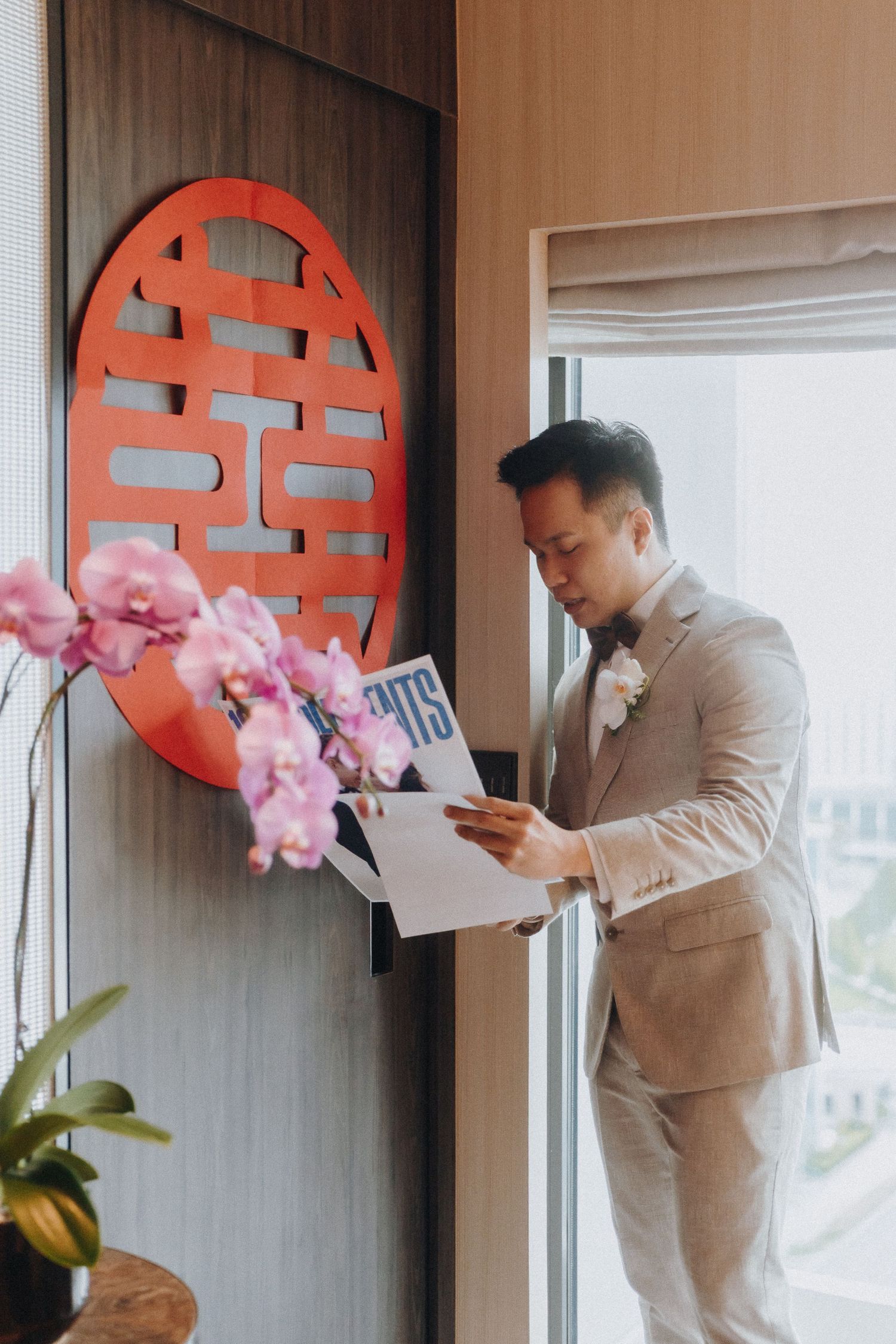 Groom in light gray suit reads letter next to red traditional Chinese wedding decorations.