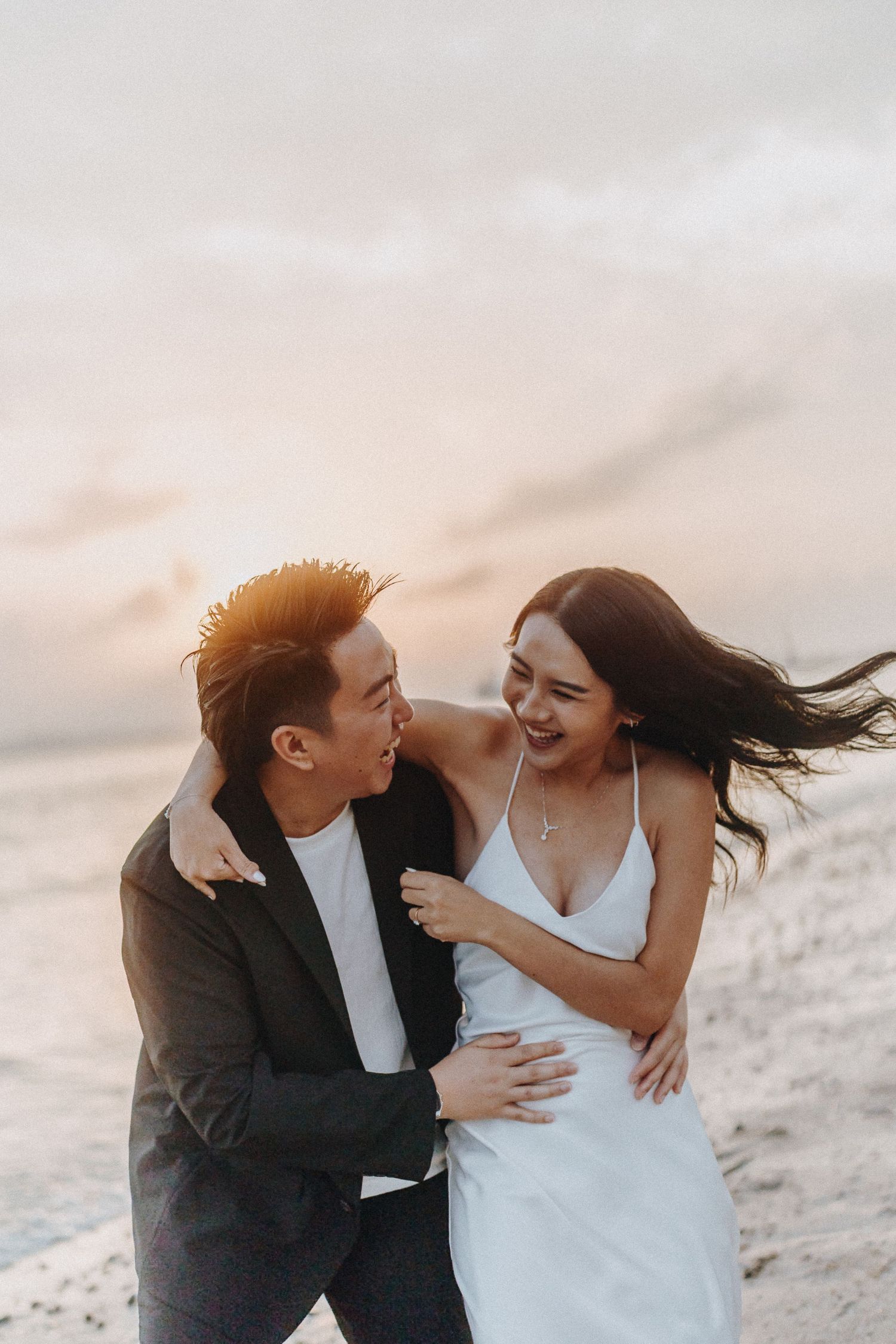 Couple in wedding attire laughing and embracing on a sandy beach at golden hour.