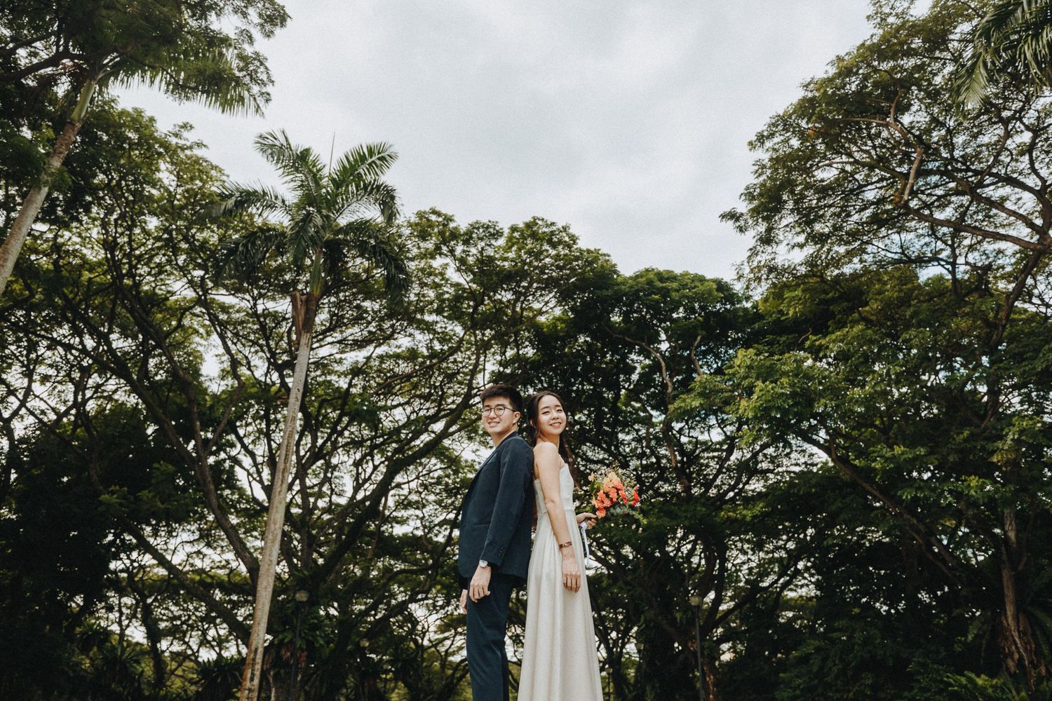 Couple poses in formal wear against a backdrop of tall green trees under an overcast sky.