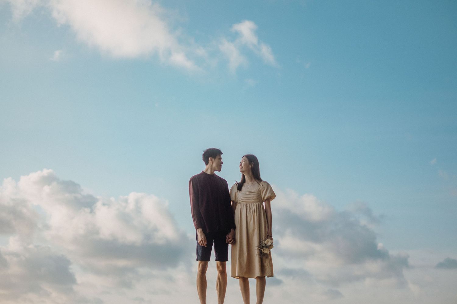 A couple shares a romantic moment on a beach at sunset with blue skies and fluffy clouds.