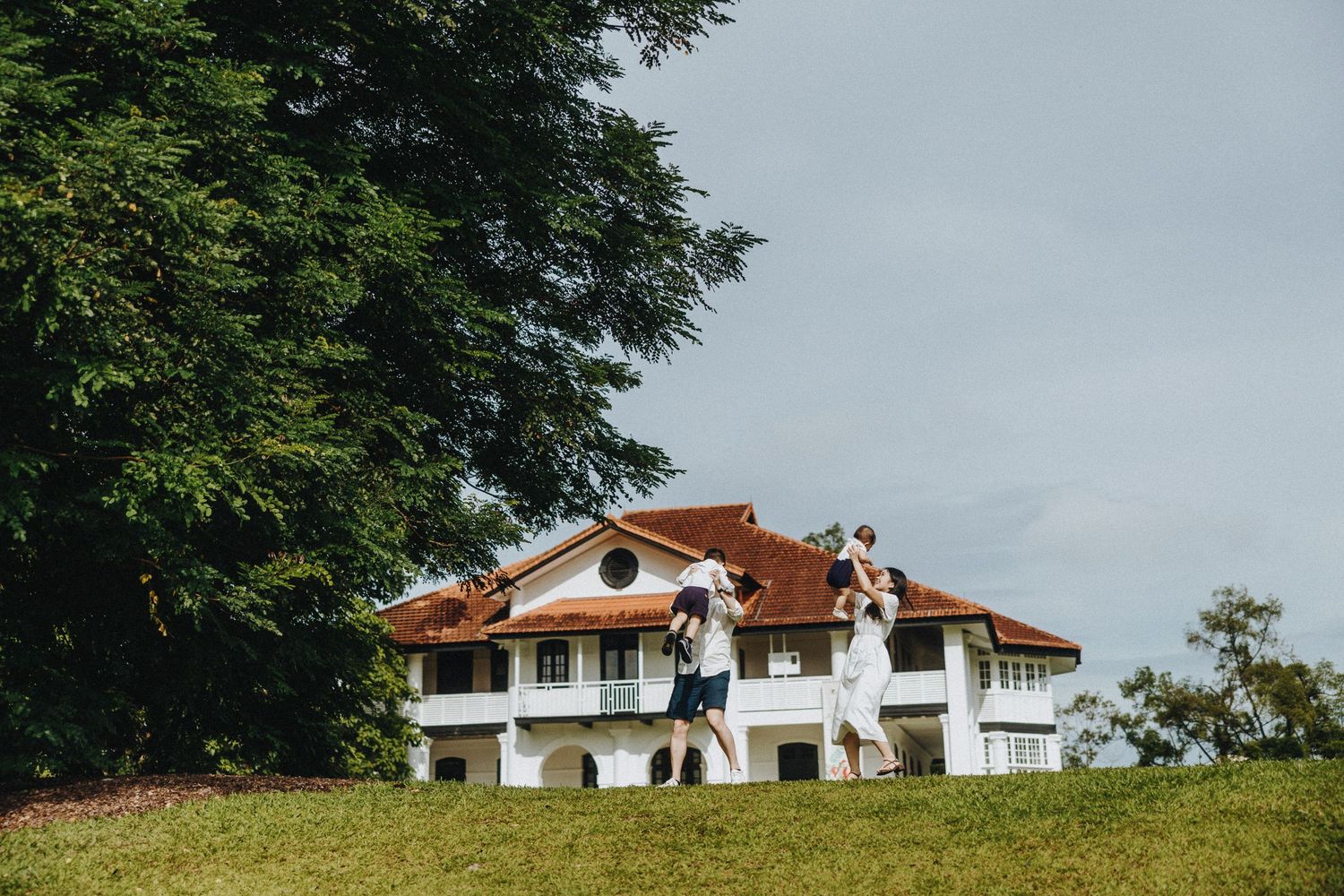 A white colonial-style mansion with red terracotta roof sits atop a grassy hillside beneath leafy green trees.