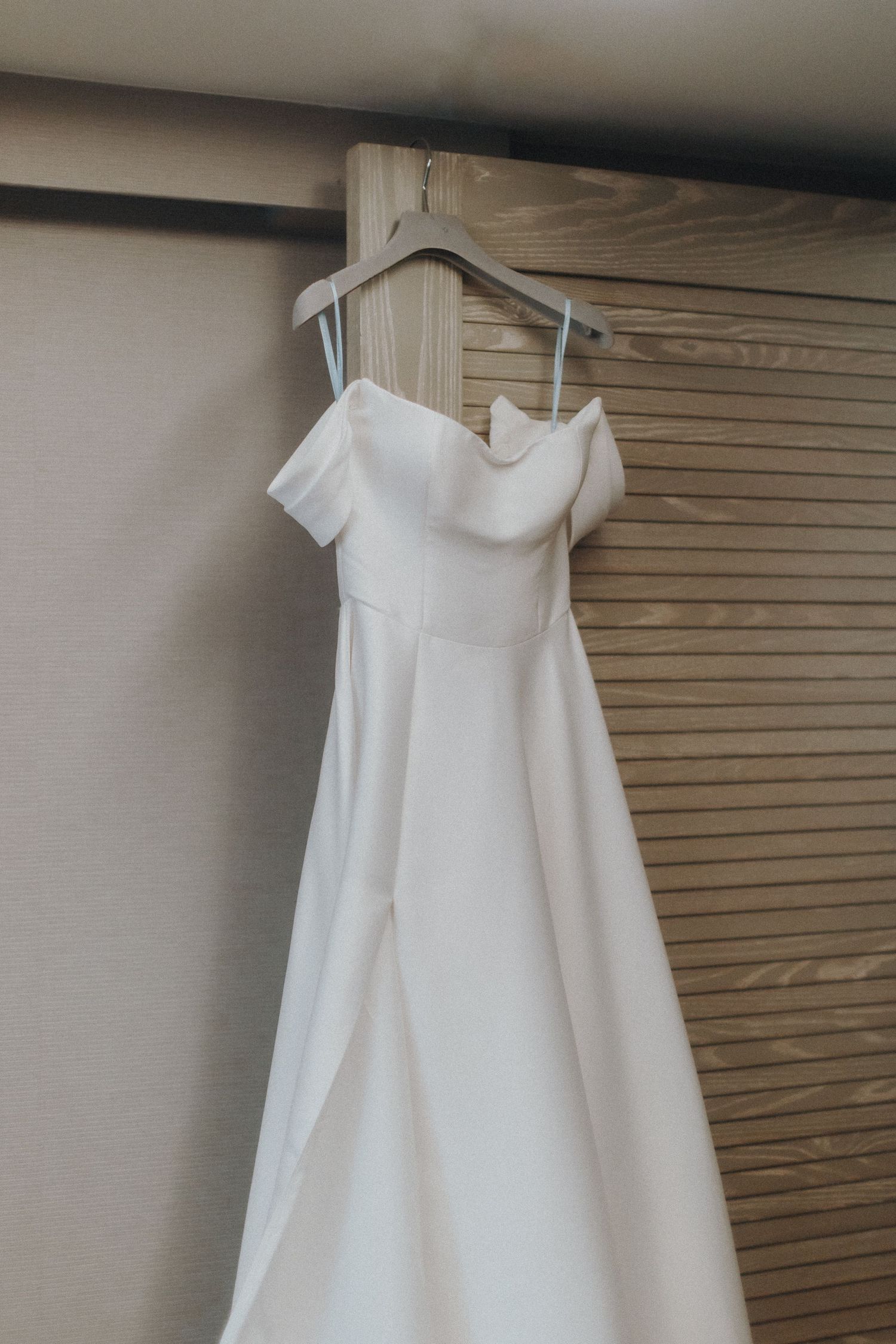An elegant white off-shoulder wedding dress hangs on a wooden hanger against a slatted wall.