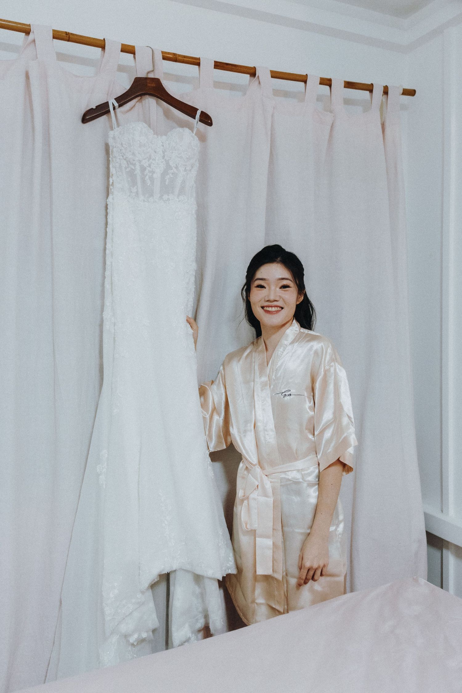 A white wedding dress hangs on display next to a person wearing a silk robe in a bridal preparation room.