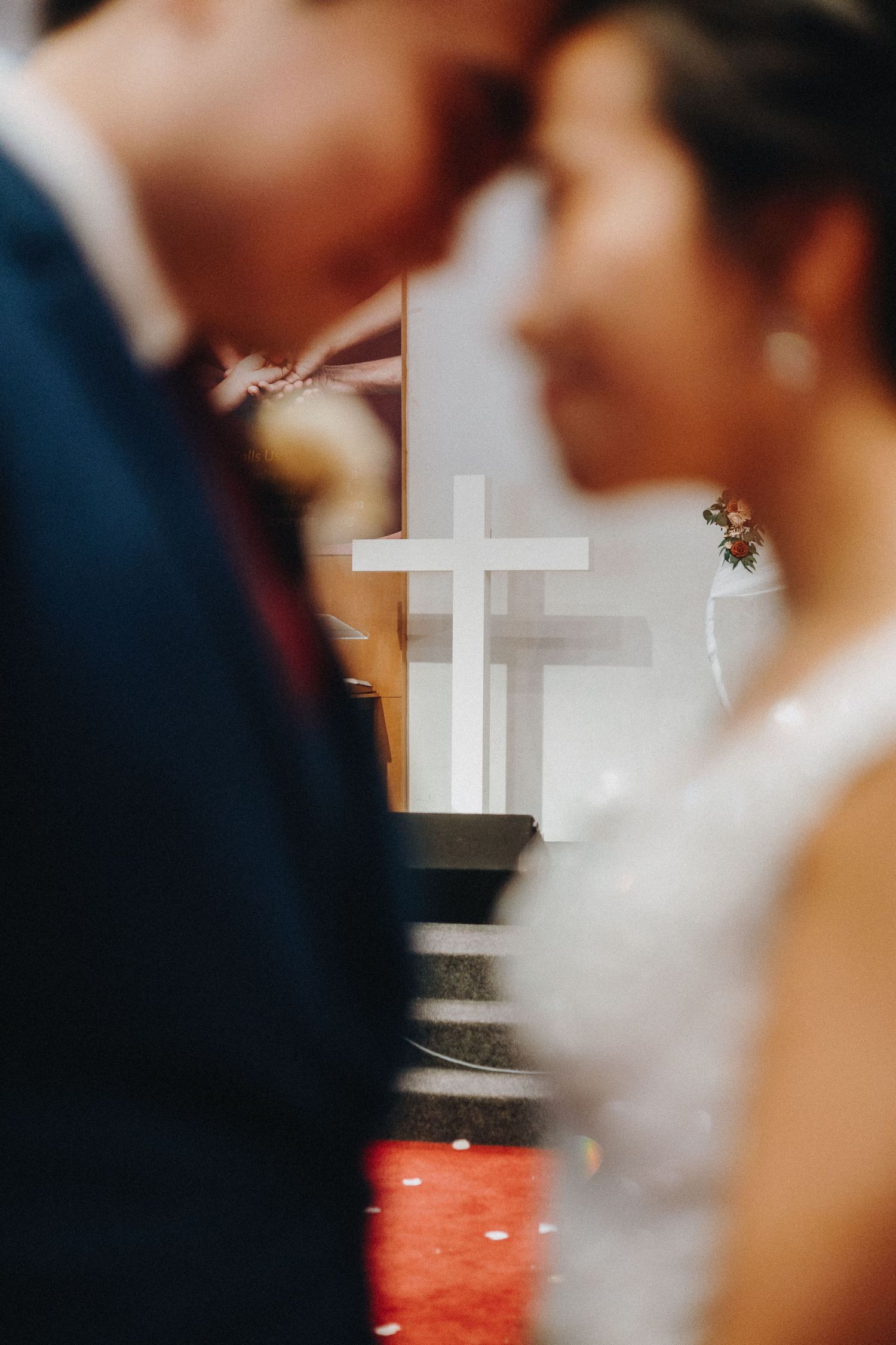 A soft-focus wedding ceremony shot with a white cross visible between a couple in the background.