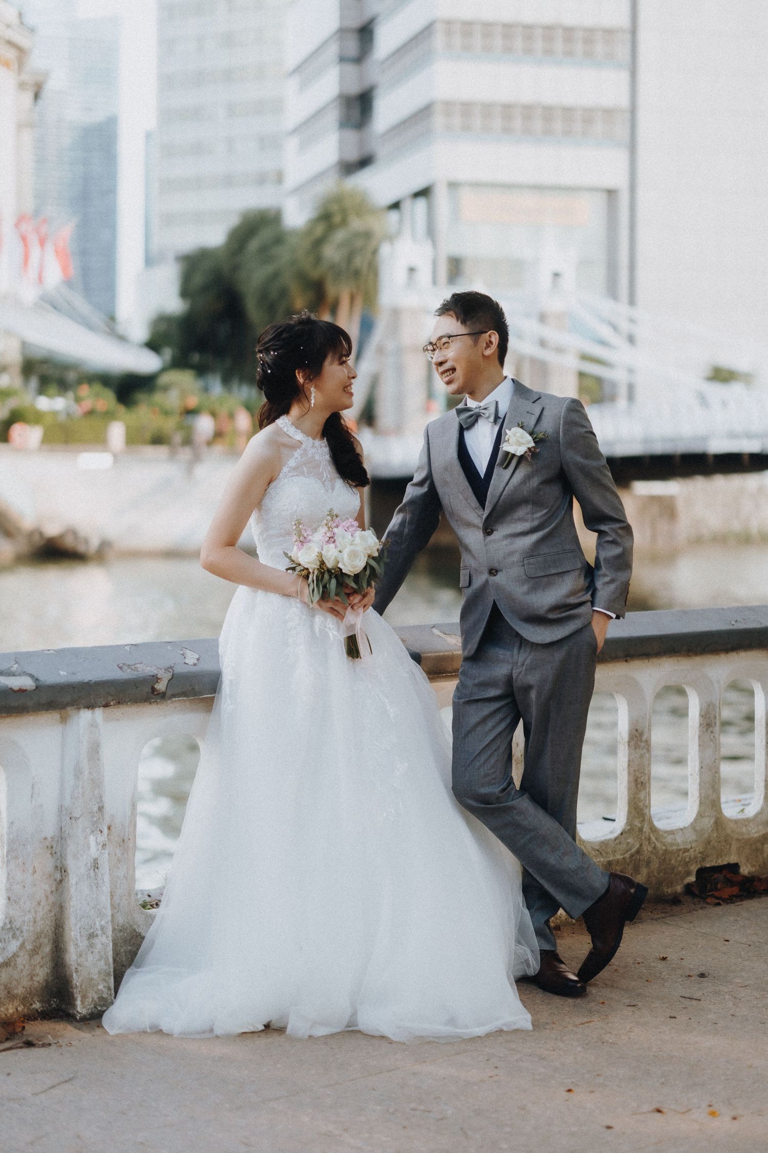 A joyful wedding couple sharing a candid moment along a waterfront with modern buildings in the background.