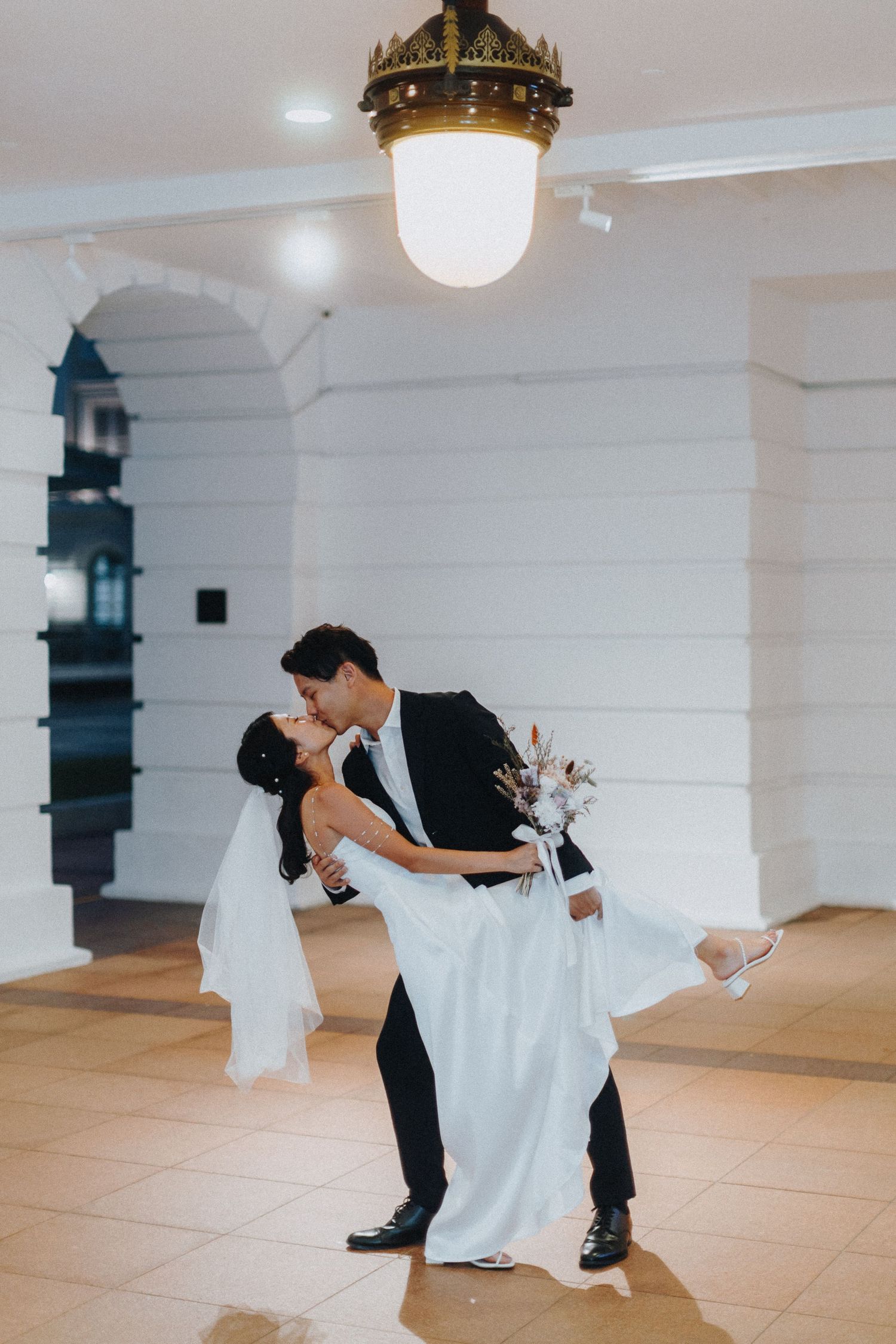 A dramatic dip during a wedding dance beneath an antique pendant light in a bright hallway.