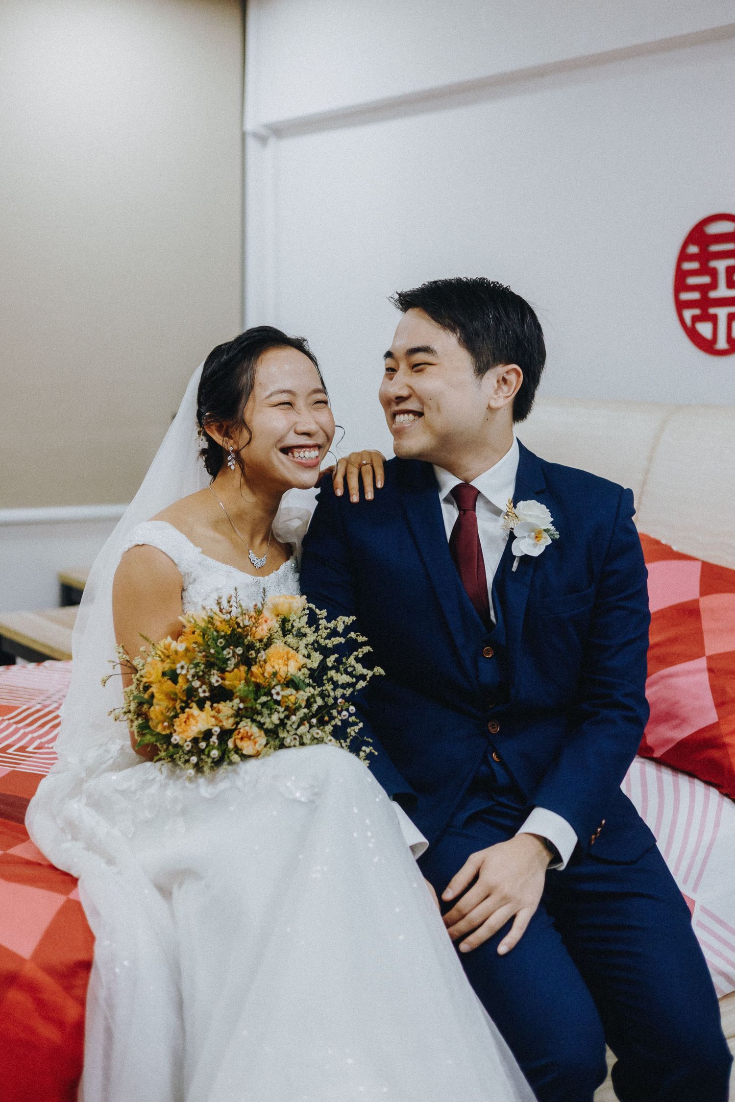 A wedding couple share a joyful moment while seated on a red couch with traditional Chinese decor.