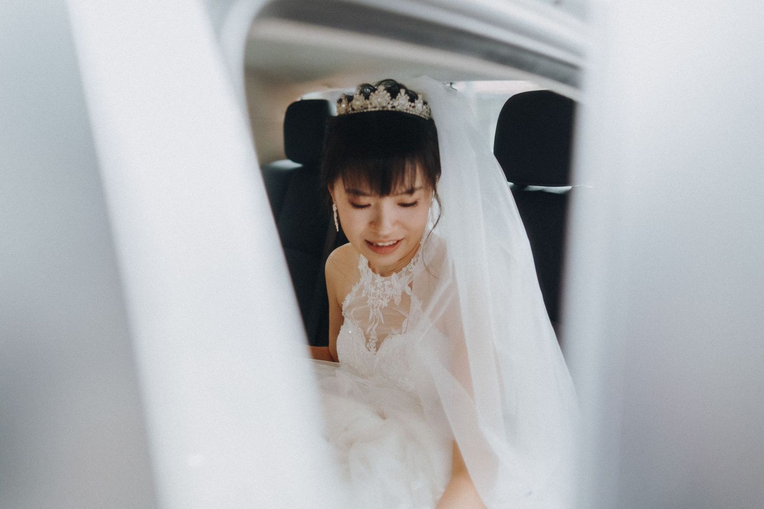 A bride smiles while sitting in the backseat of a car wearing a delicate veil and tiara.
