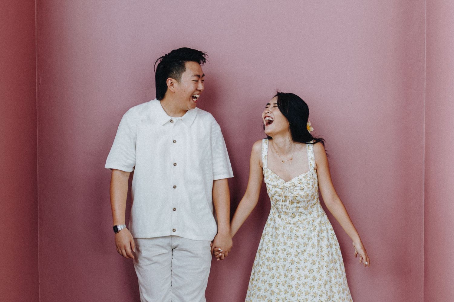 Couple shares a joyful moment against a pink wall in matching white outfits.